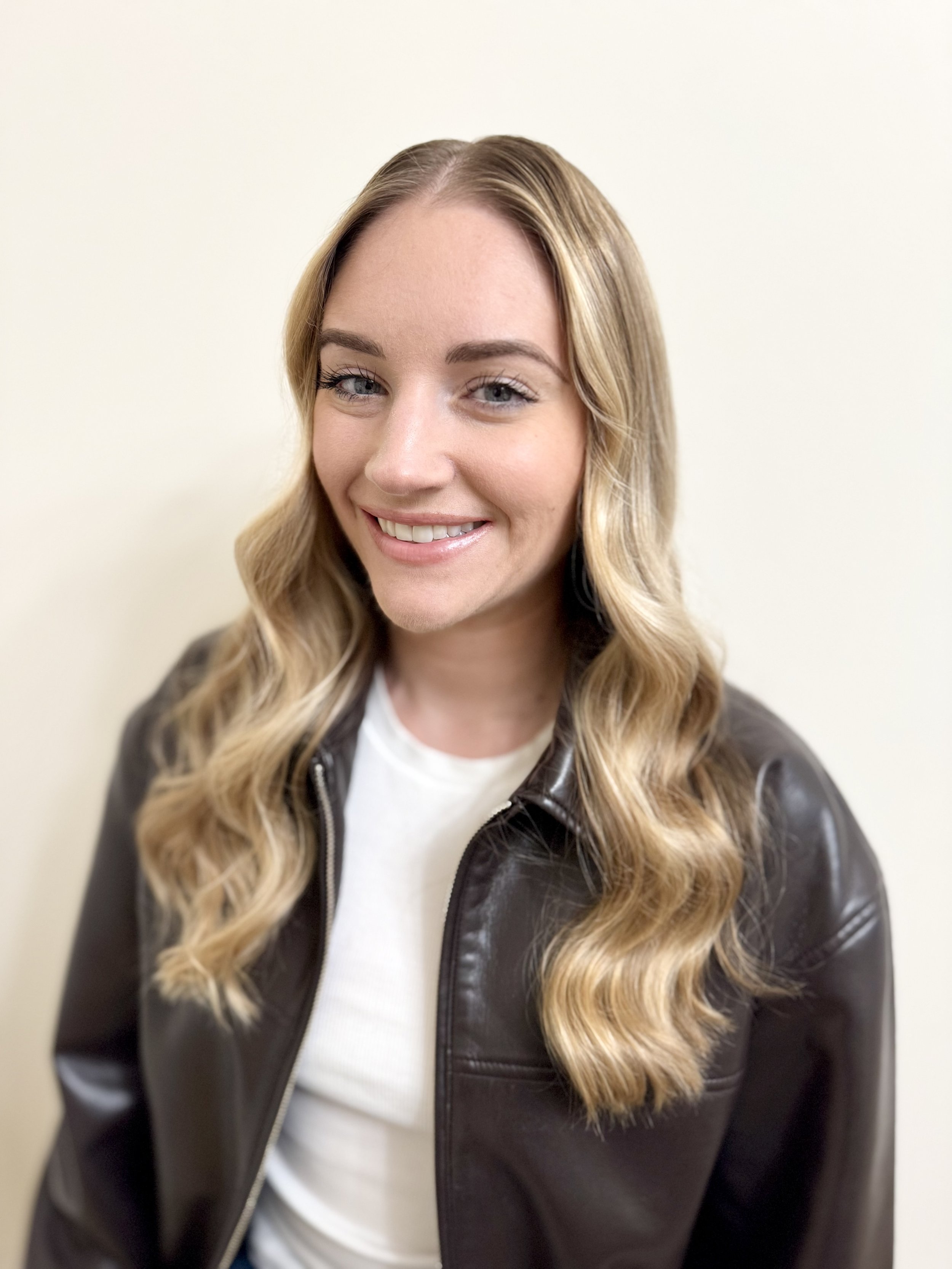 A smiling woman with long, wavy blonde hair, wearing a black leather jacket over a white shirt, standing against a plain light-colored wall.