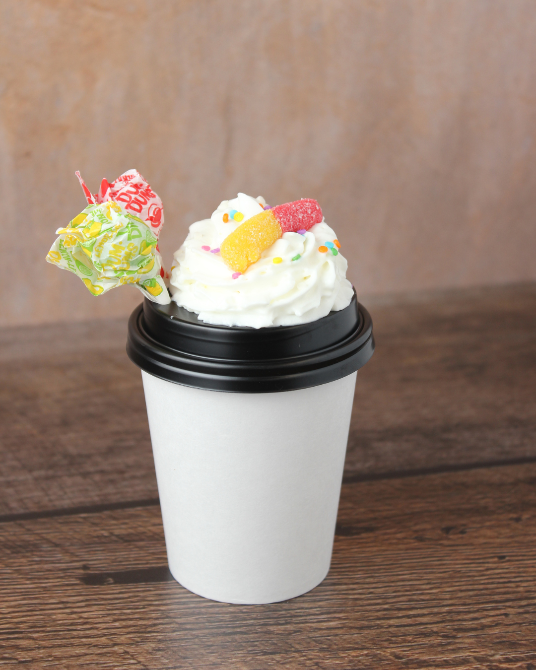 Paper coffee cup with whipped cream, colorful sprinkles, gummy candy, and decorative lollipop on top, placed on a wooden surface.