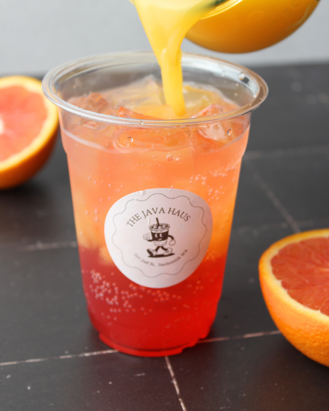 A clear plastic cup filled with pink grapefruit soda with ice cubes, with a logo reading 'The Java Haus' on the front. Whole grapefruits are around the cup on a dark surface.