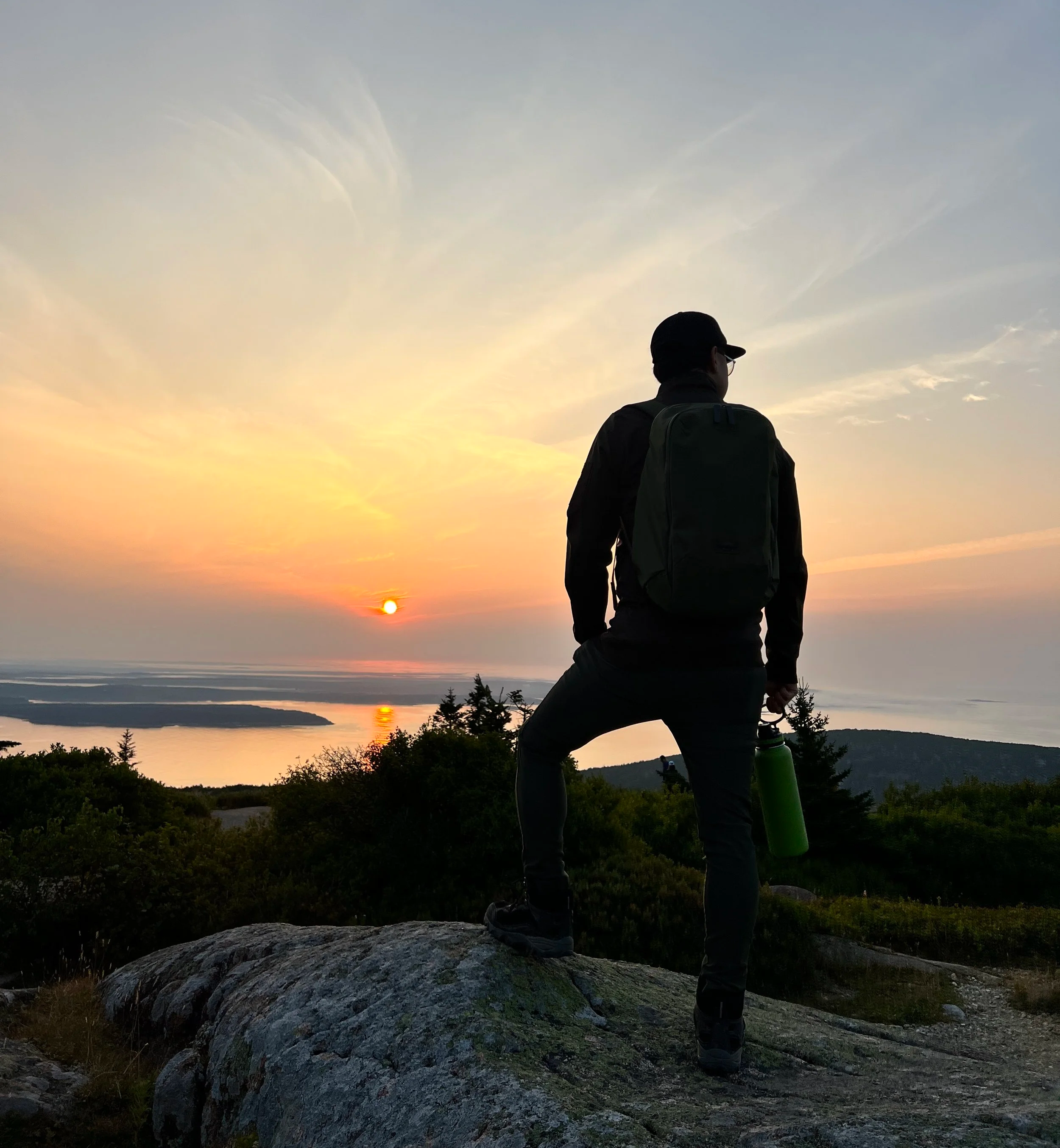 Silhouette of a person standing on a rock during sunset, overlooking trees, water, and a distant horizon.