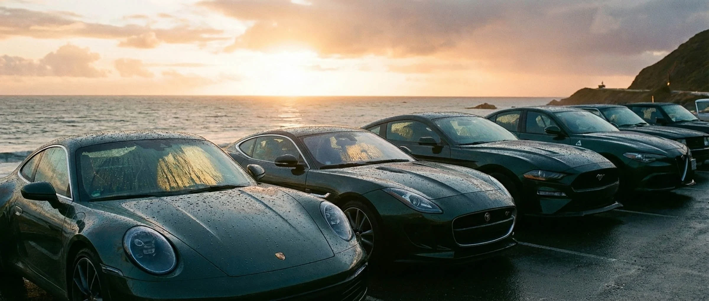 A row of parked cars along a beachside during sunset, with hills in the background and wet windows on the cars reflecting the sunset sky.