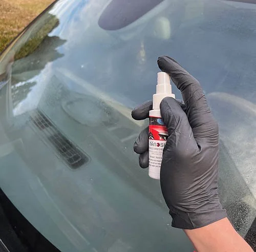 Person wearing black gloves holding a small spray bottle near a car windshield.