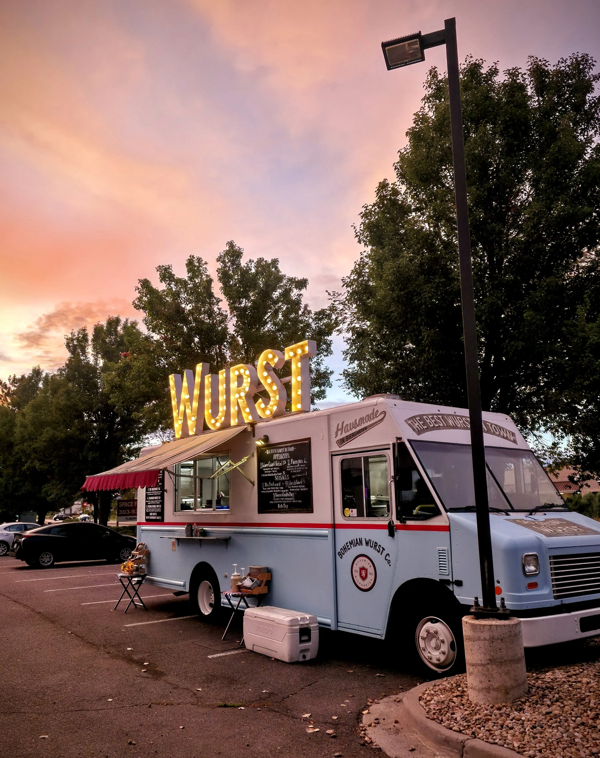 A light blue vintage food truck with a large illuminated sign spelling 'WURST' on top, parked in a lot during sunset with pink and purple skies, trees, and other cars in the background.