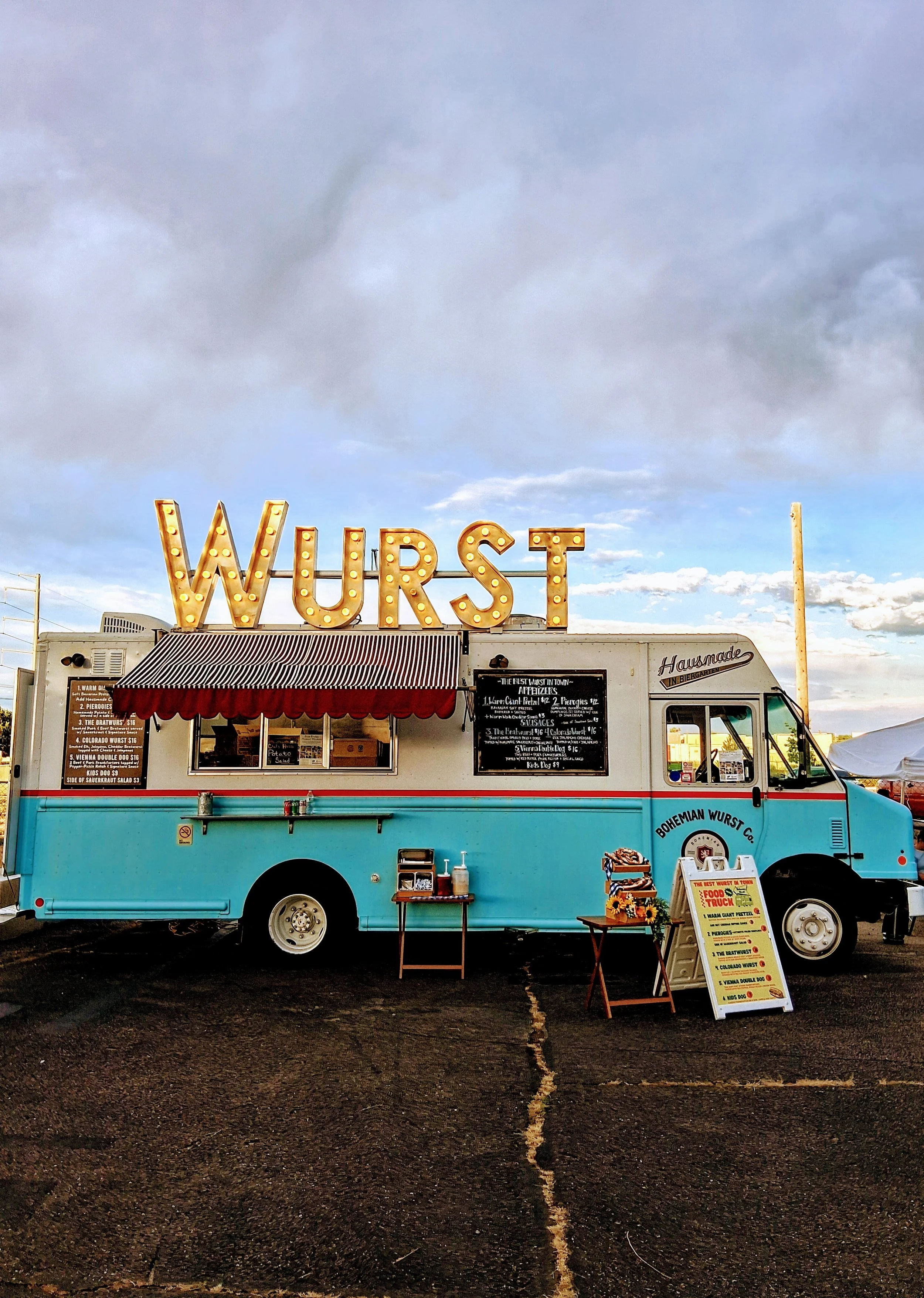 Colorful food truck with a large illuminated 'WURST' sign on top, serving sausages and deli items, with a menu board outside in a parking lot during the day under a sky with clouds.