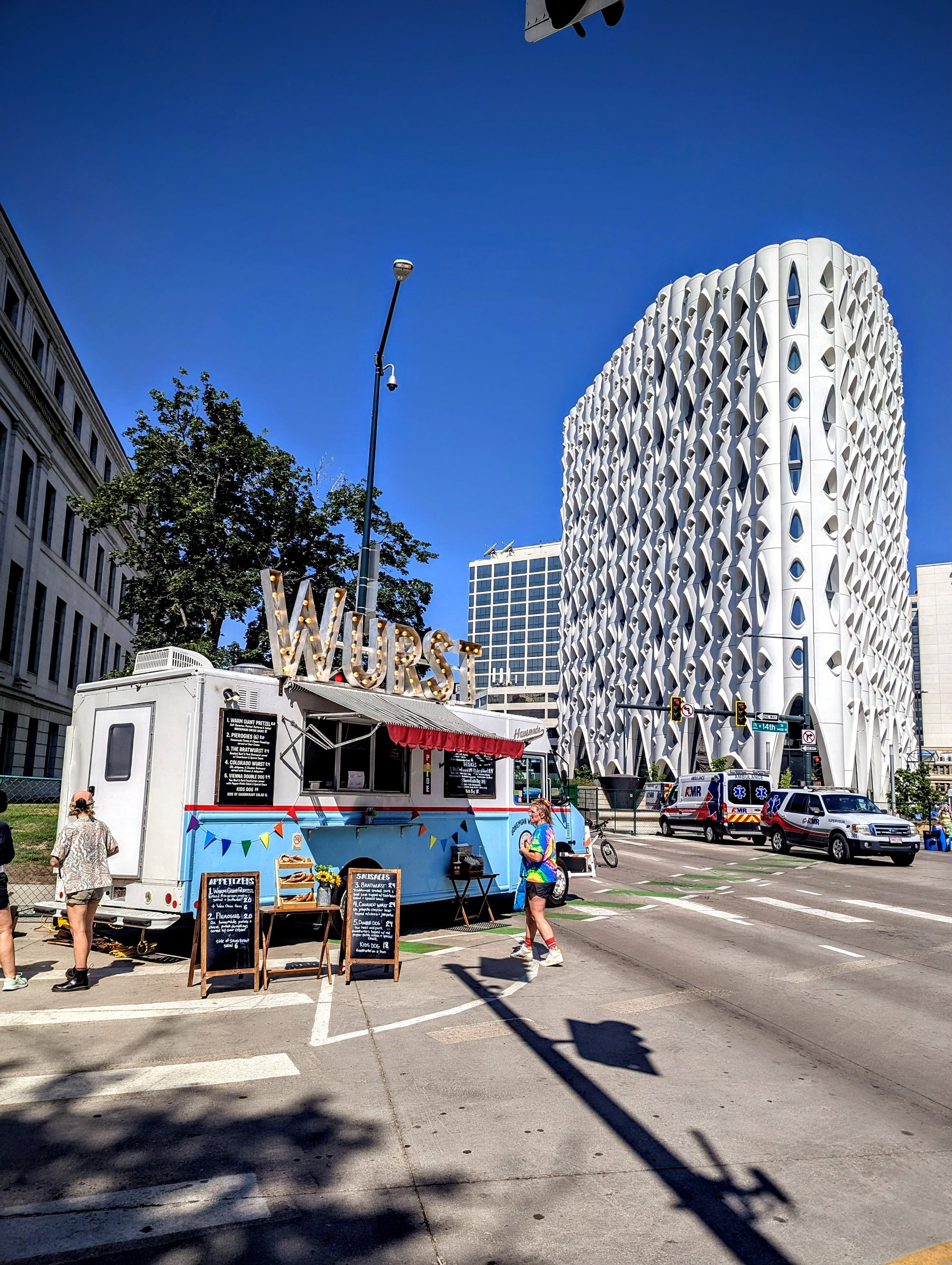 Street scene with food truck named 'WURST,' people walking, modern white building with an intricate facade, emergency vehicles, and traffic lights on a sunny day.