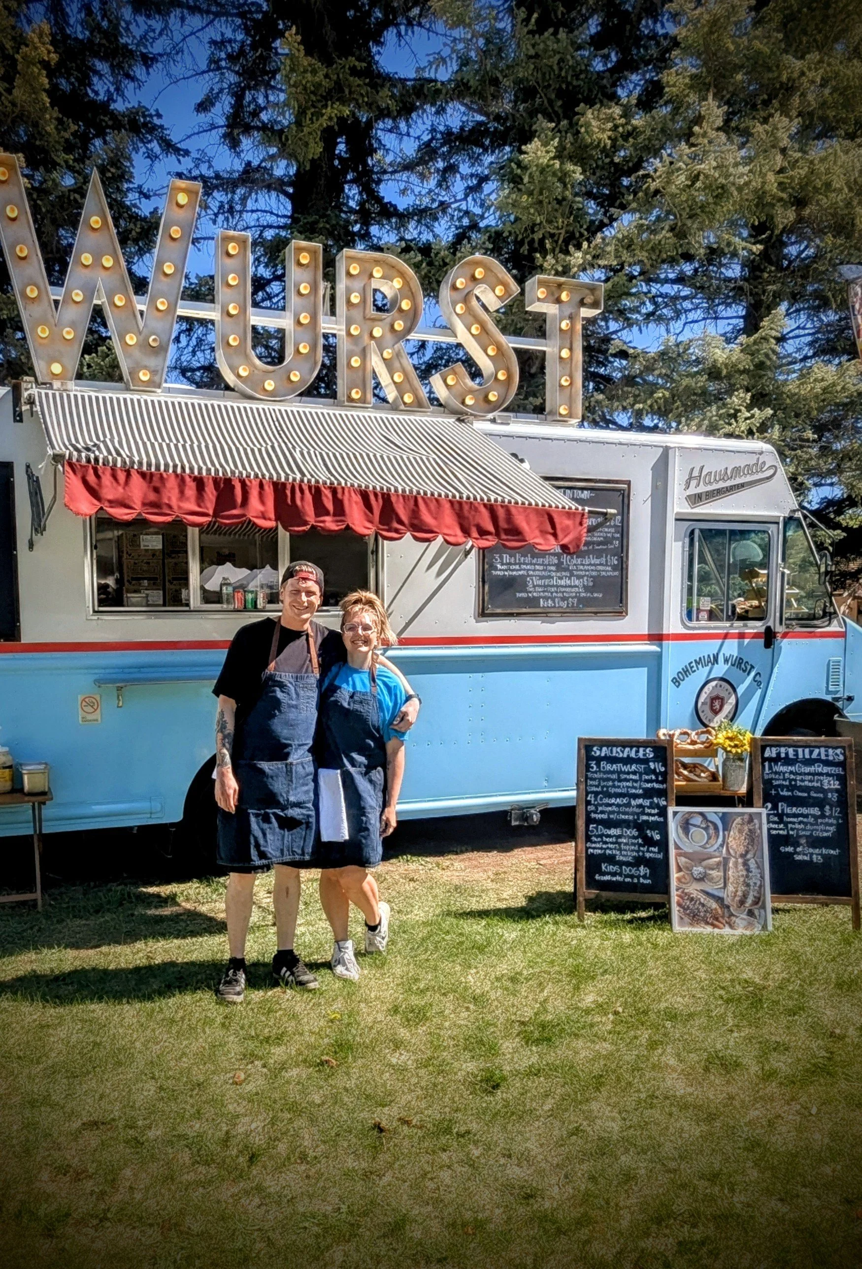 Two smiling food vendors, a man and a woman, standing in front of a colorful food truck with a large illuminated "WURST" sign on top, outdoor setting with trees and grass.