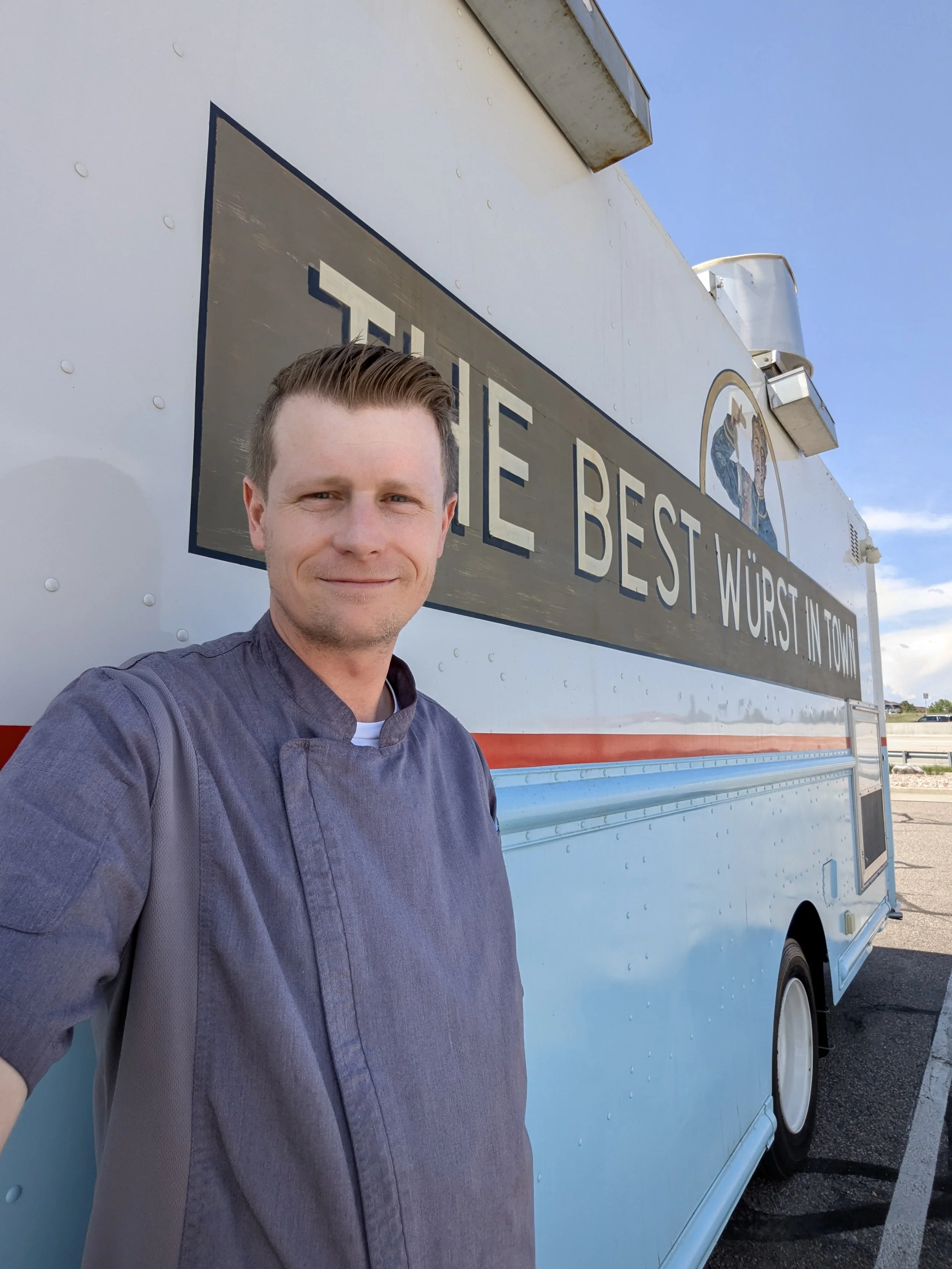 A man in a chef's coat standing in front of a food truck with the sign 'The Best Wurst in Town' under a blue sky.