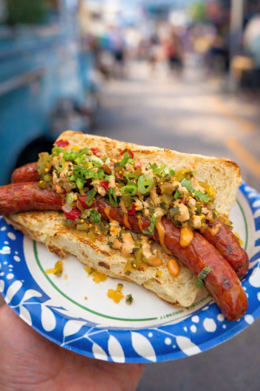 A hot dog in a bun topped with chopped green onions, relish, and spicy mustard, held in front of a blurred street scene.