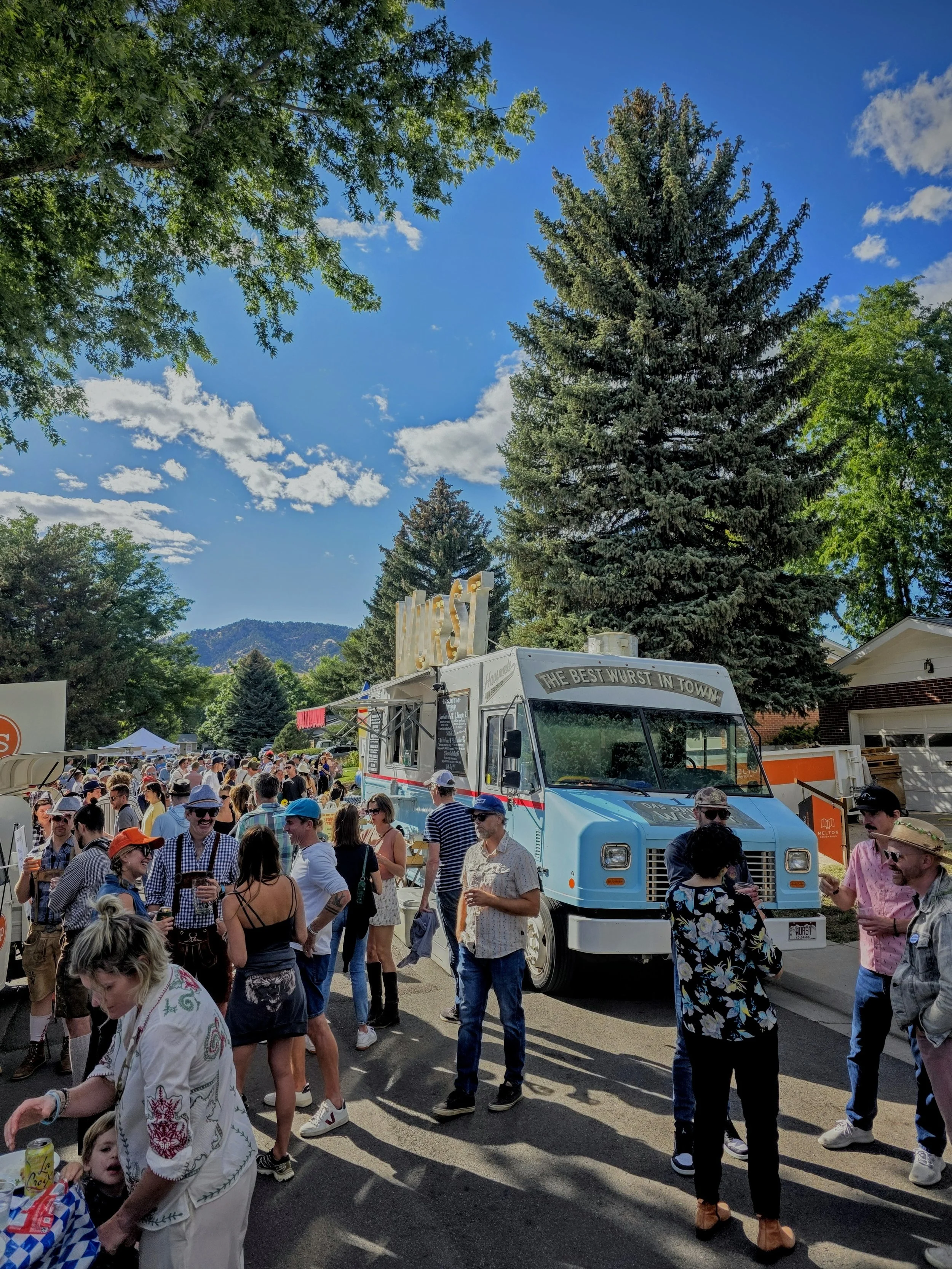 Crowd of people gathered around a blue food truck at an outdoor festival on a sunny day with trees and mountains in the background.