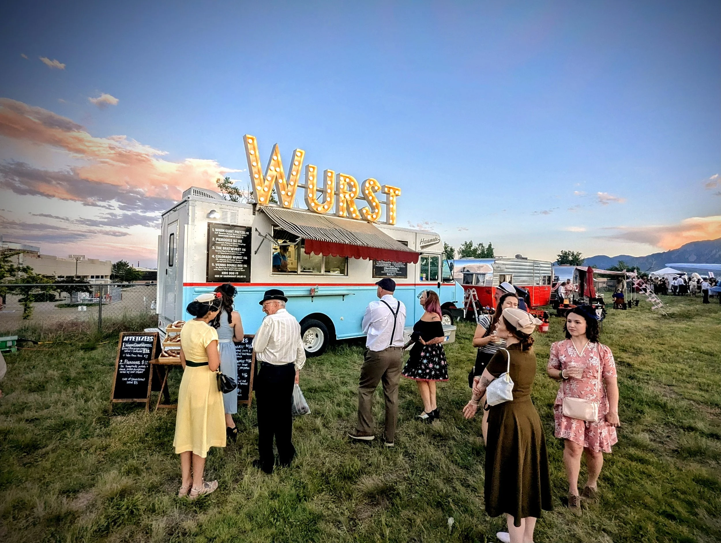 A vintage food truck named 'Wurst' serving at an outdoor event with people waiting and socializing on a grassy field during sunset.