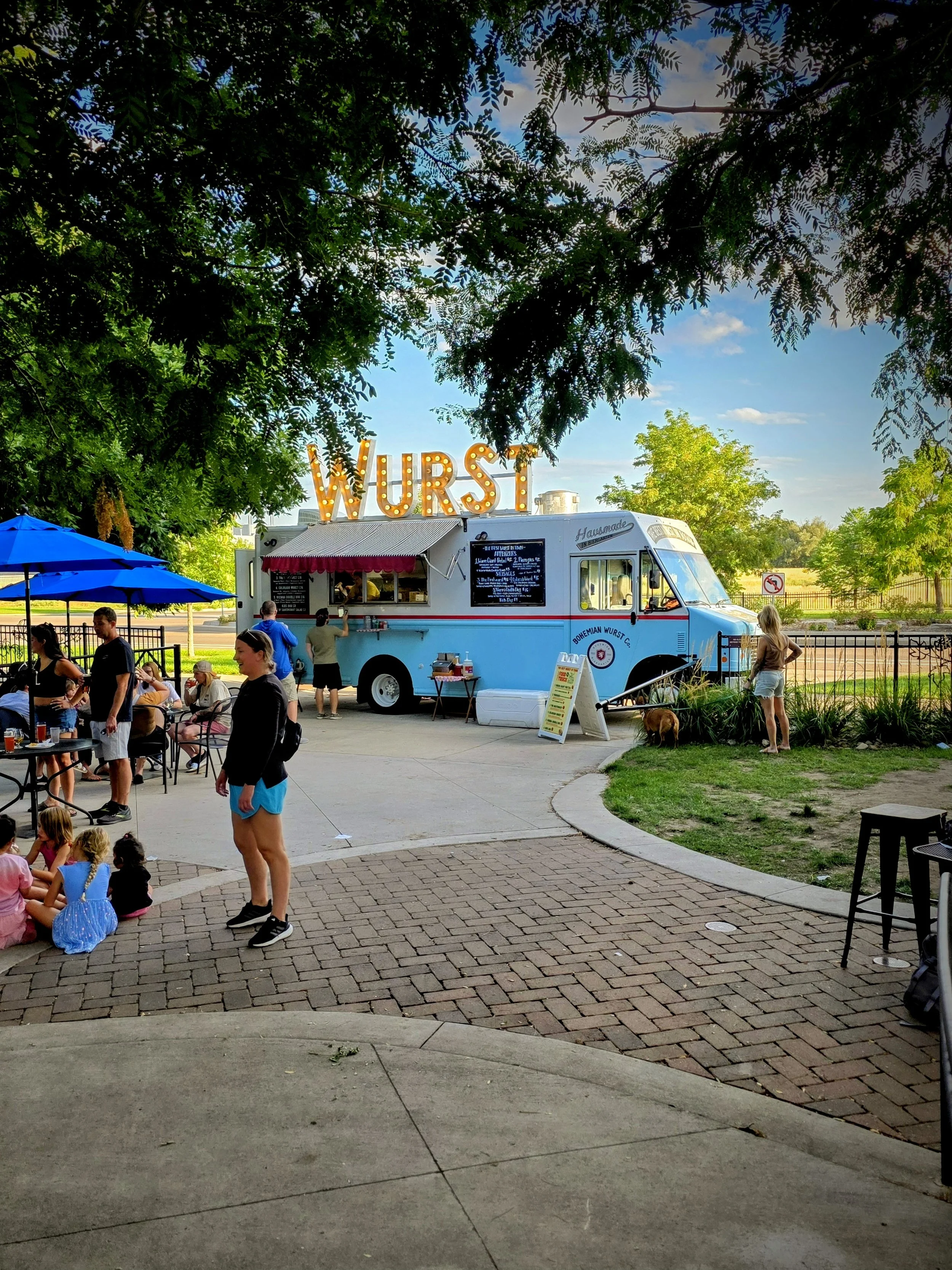 Food truck with large illuminated 'WURST' sign, people gathered around outdoors on a sunny day, some sitting at tables, children sitting on the ground, and a woman with a dog nearby.