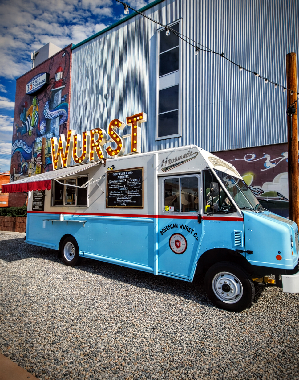 A vintage food truck painted light blue with red and white accents, parked on gravel in front of a building with a colorful mural and large illuminated "WURST" sign.