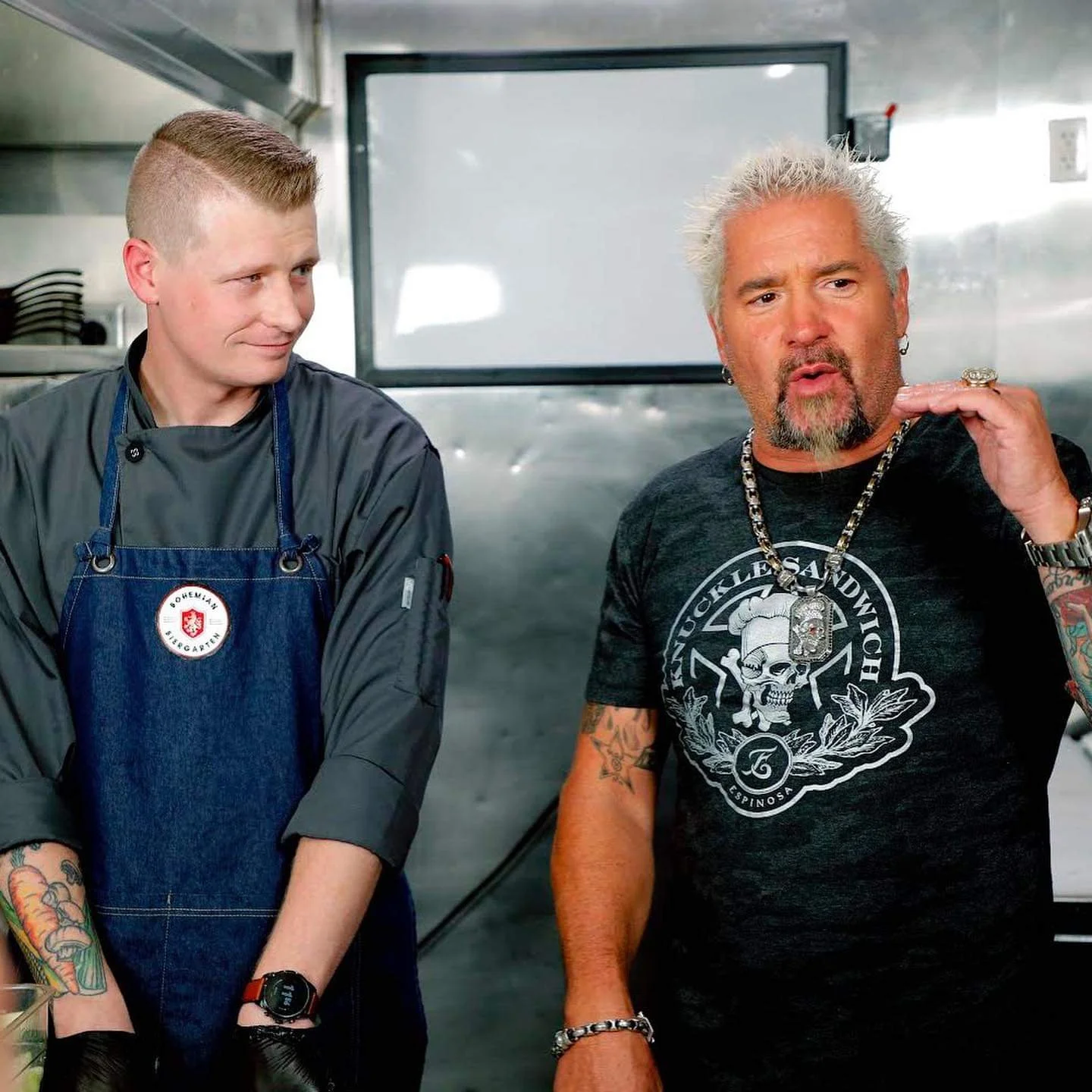 Two men inside a kitchen or food truck. The man on the left is a young chef wearing a gray chef coat and a denim apron, looking at the man on the right with a smile. The man on the right has gray hair, a beard, and tattoos, wearing a black T-shirt wi