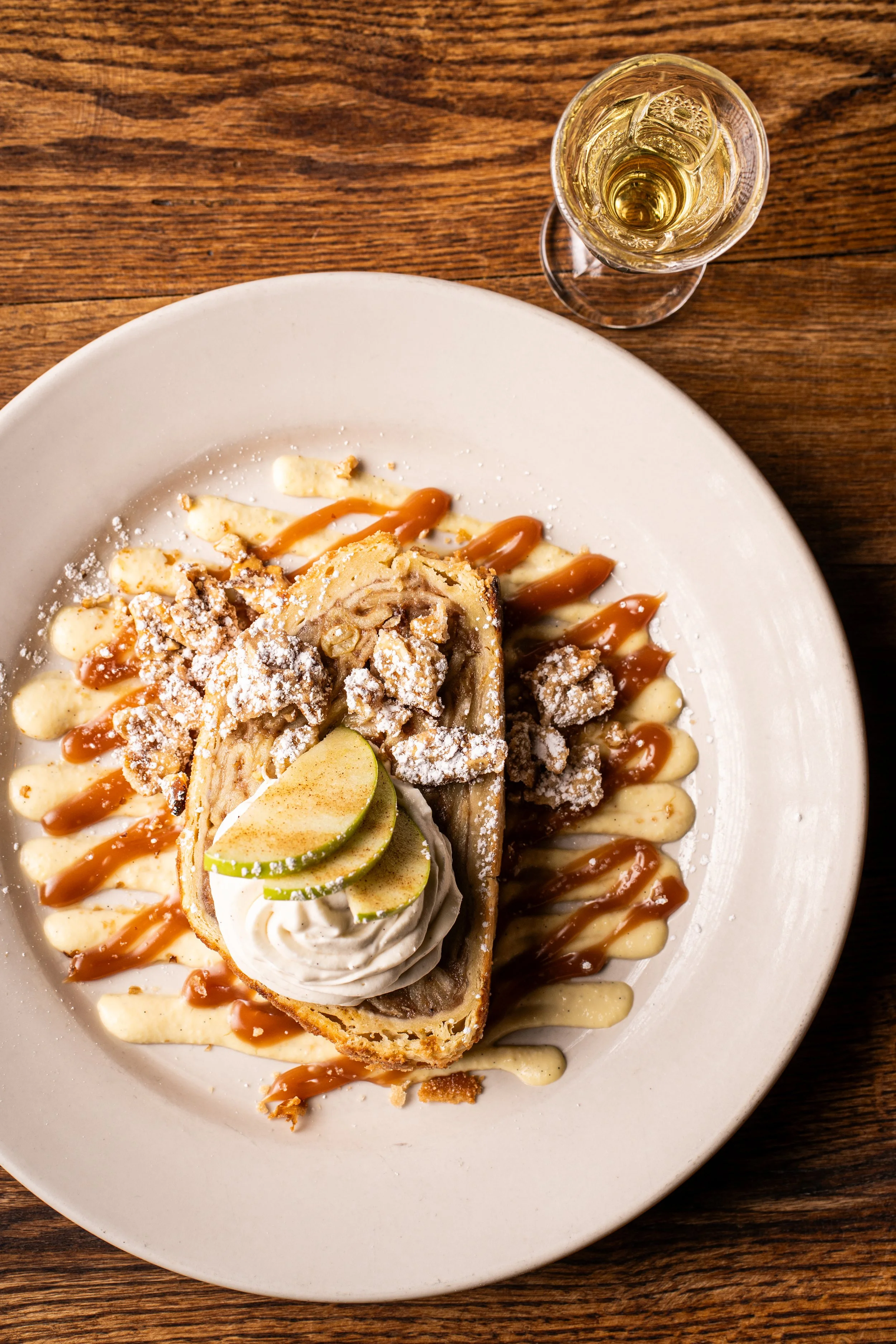 A dessert featuring a slice of apple pie topped with whipped cream and lime slices, drizzled with caramel and vanilla sauces, served on a white plate, with a glass of white wine on a wooden table.