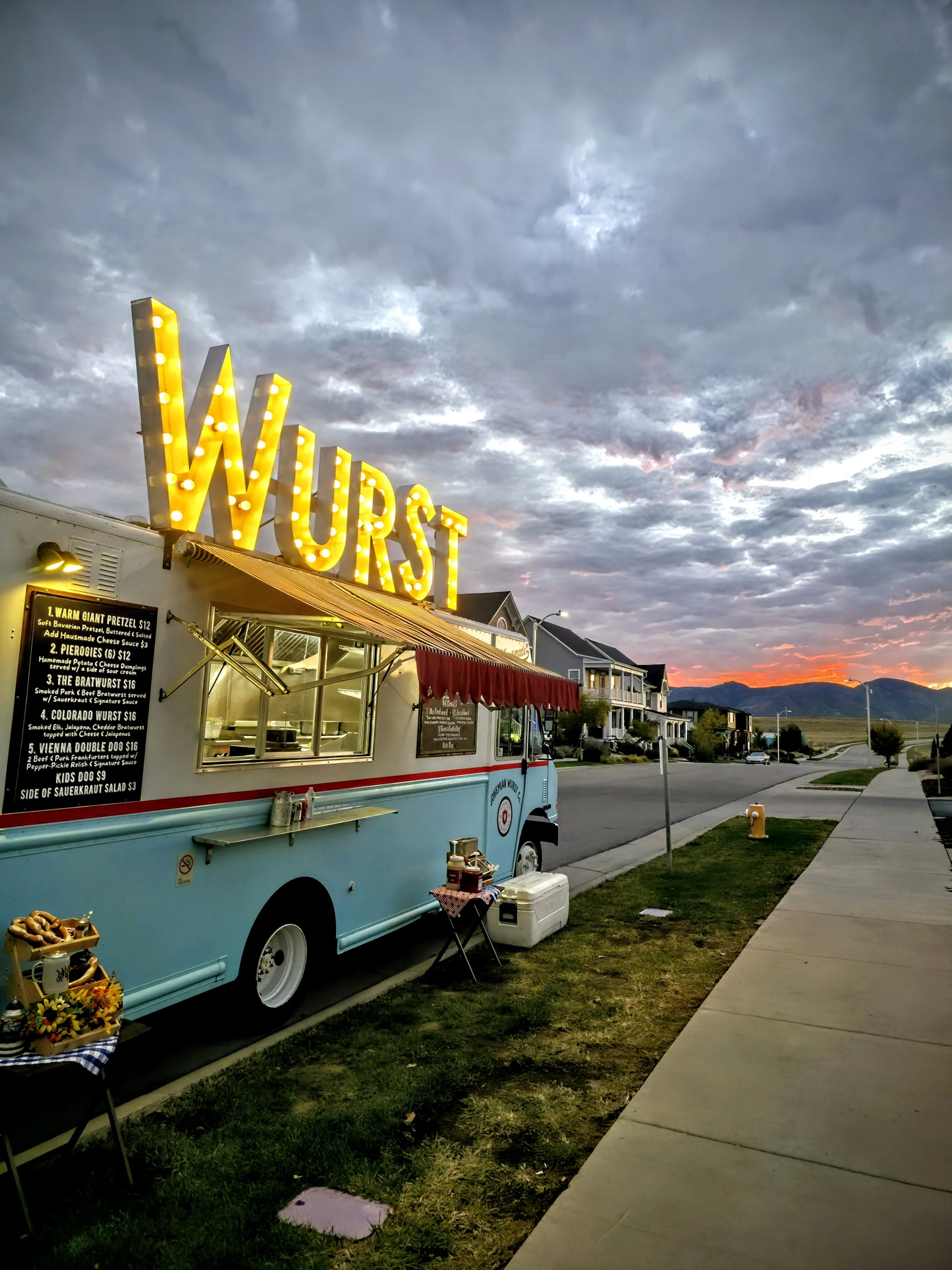 A food truck named 'Wurst' with a bright illuminated sign, parked on a street during sunset, with houses in the background and a cloudy sky overhead.