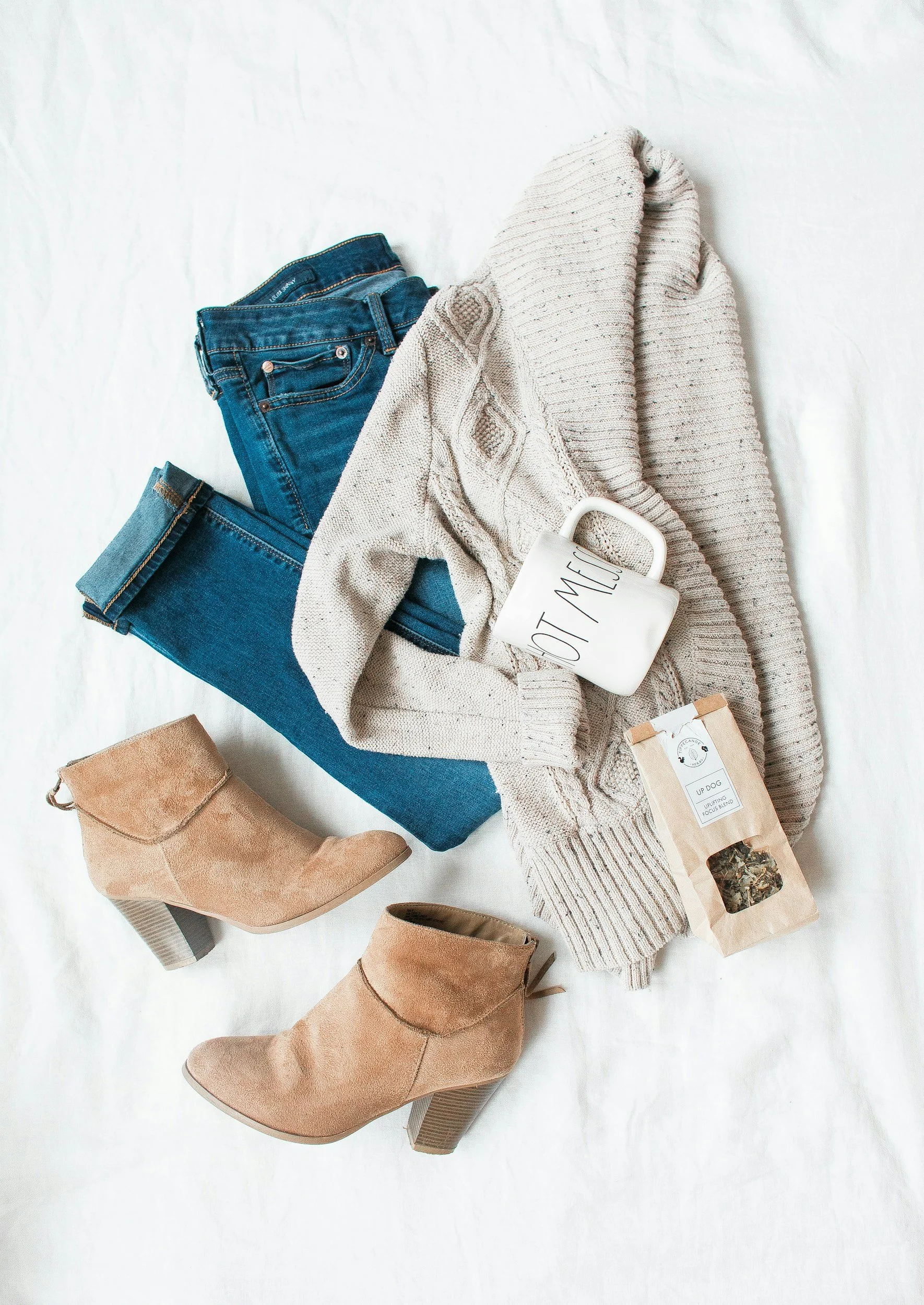 A flat lay of casual fall outfit items including a beige sweater, blue jeans, tan ankle boots, a white mug with text, and a small bag of herbal tea on a white surface.