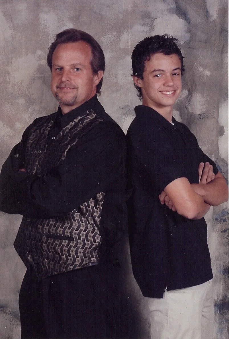 A man and a teenage boy stand back to back with arms crossed and smiling at the camera against a textured gray background.