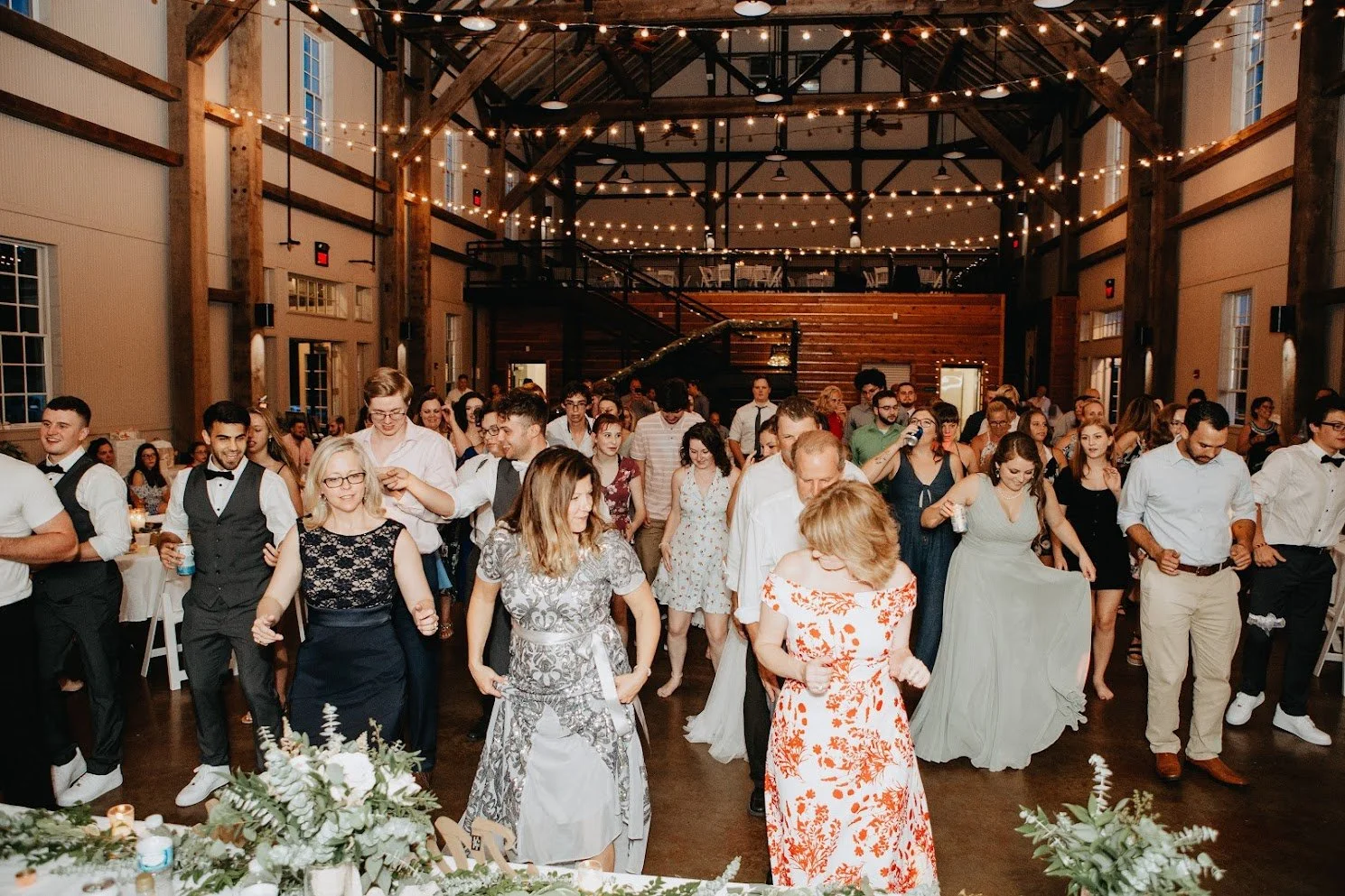 People dancing at a wedding reception in a rustic indoor venue with string lights and wooden beams.