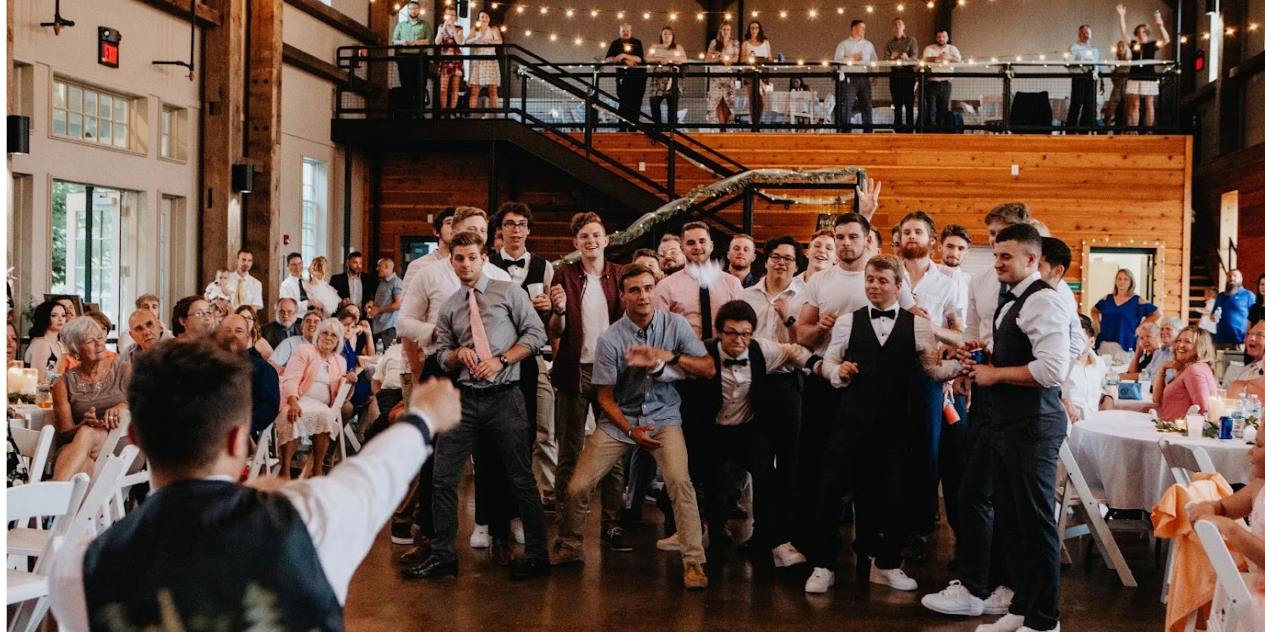 A wedding reception with a large group of people, some dancers in the foreground, and guests seated at tables in the background inside a rustic venue with wooden walls and a high ceiling.