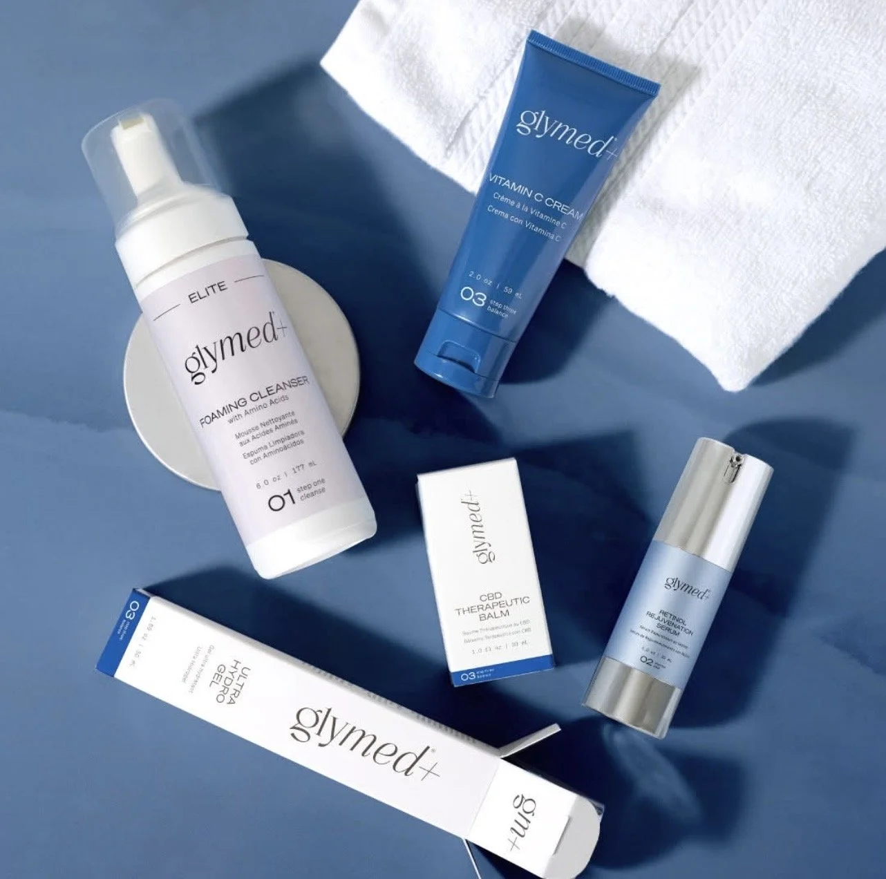 GlyMed+ skincare products, including foaming cleanser, vitamin C cream, therapeutic balm, rejuvenating serum, and eye cream, arranged on a blue surface.