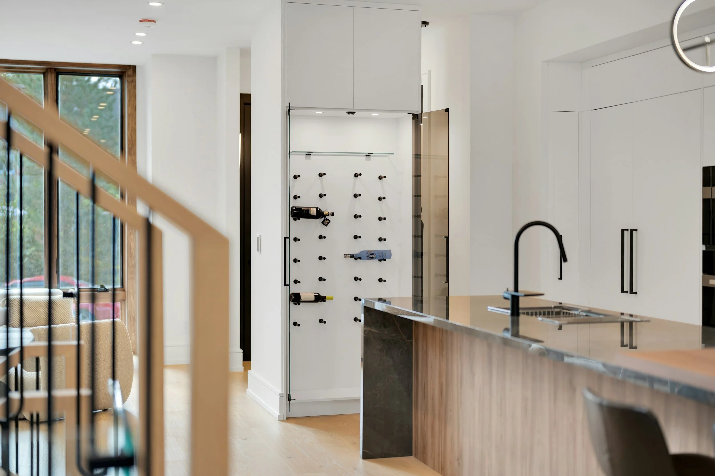 Modern kitchen with a white island, black faucet, and a white wall with cabinets. A wine rack is visible on a panel on the wall next to the cabinets, with several wine bottles placed horizontally.