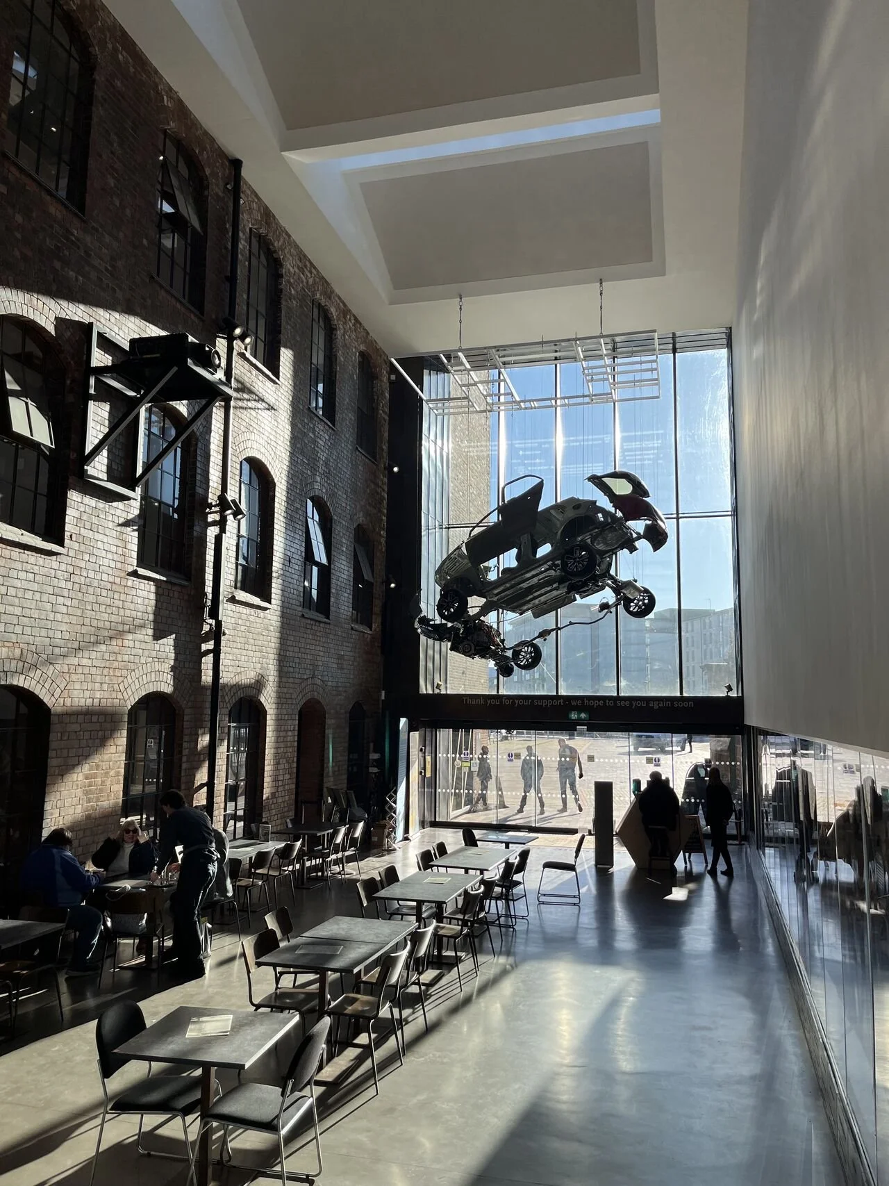 Interior of a modern coffee shop with a brick wall on the left and large glass windows at the front. There is an art installation hanging from the ceiling resembling cars. Several people are sitting at tables, and sunlight is streaming in through the windows.