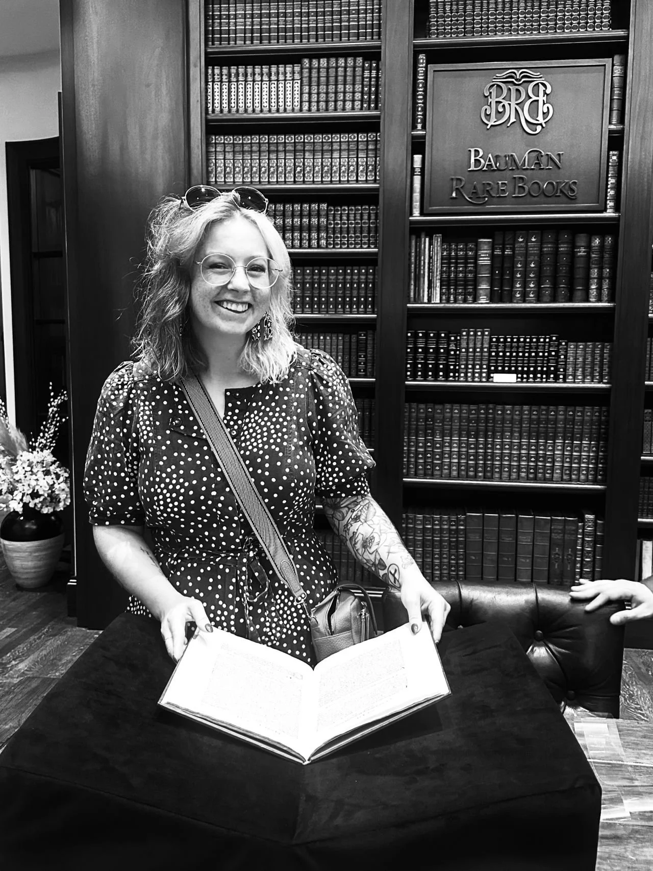 A black and white image of a woman stood holding an open book, resting on a table. There is a bookcase behind her.