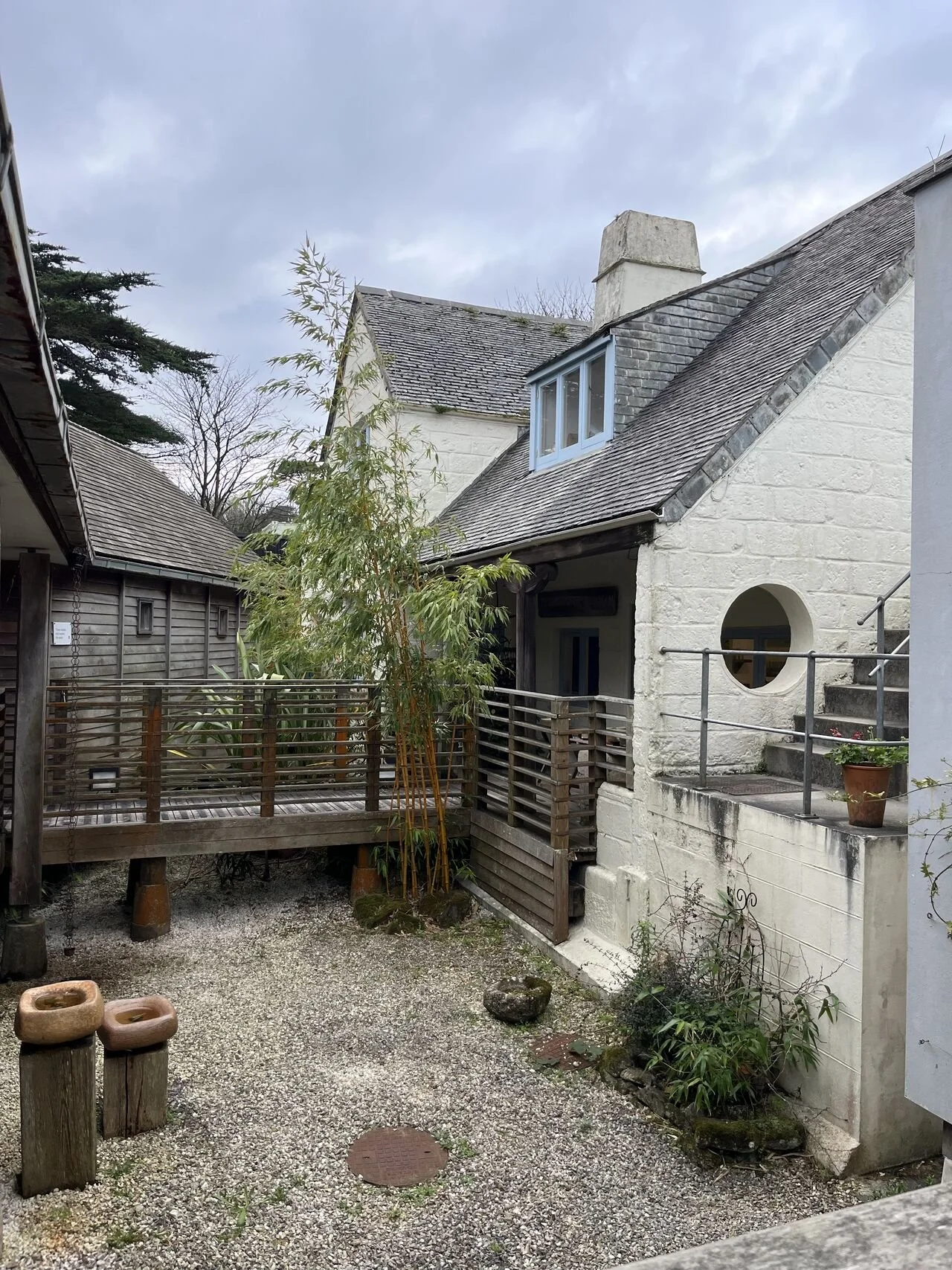A small outdoor courtyard with gravel ground, a wooden deck, potted plants, and a white brick house with a grey, tilted roof and a chimney. Overcast sky.
