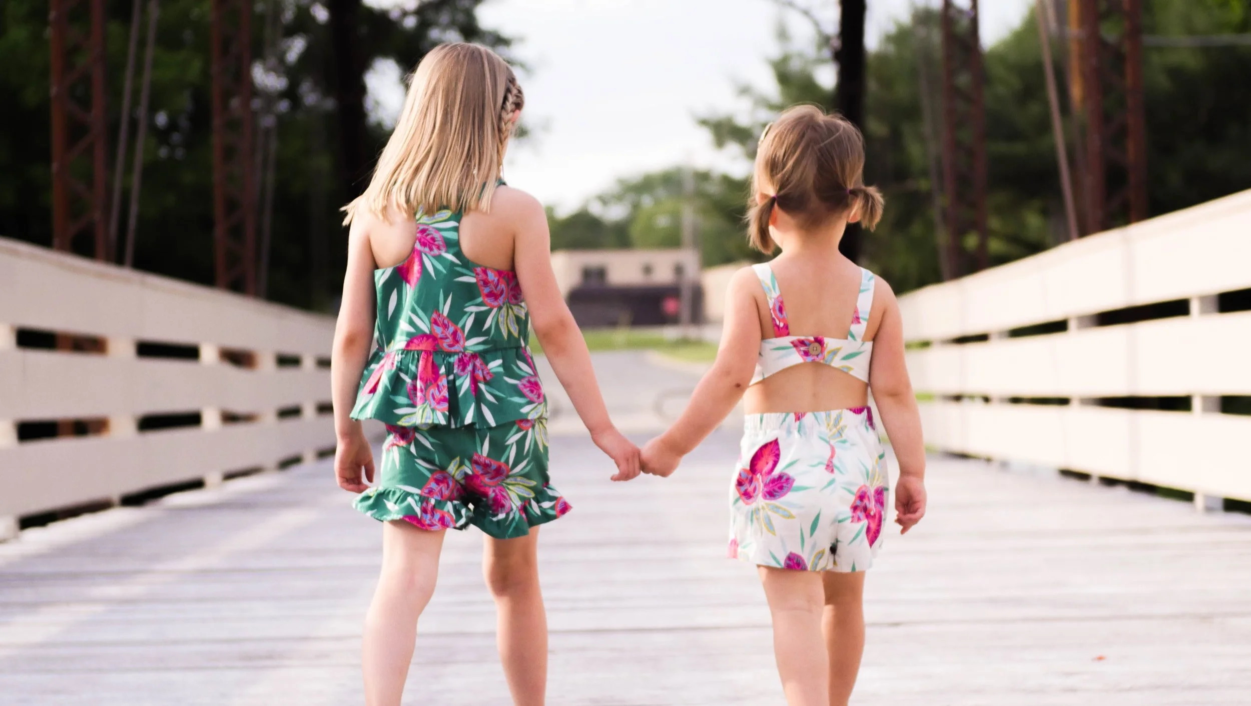 Two young girls wearing matching floral dresses holding hands and walking on a wooden bridge with trees in the background.