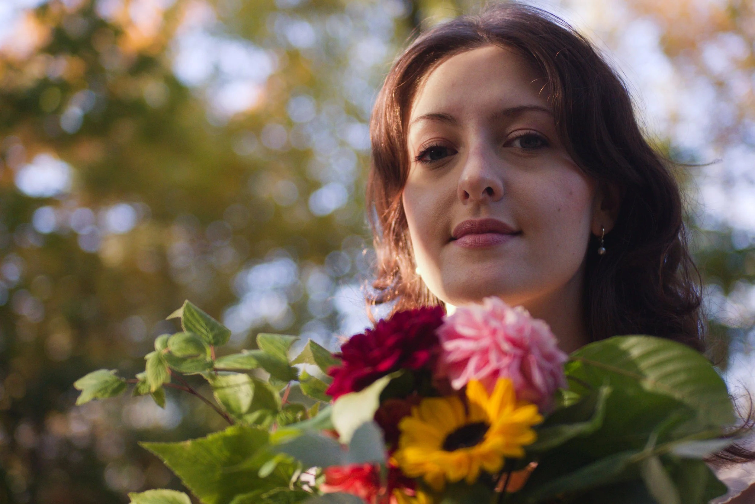 Close-up of a woman with dark hair and light skin, holding a bouquet of colorful flowers, standing outdoors with blurred trees and sunlight in the background.