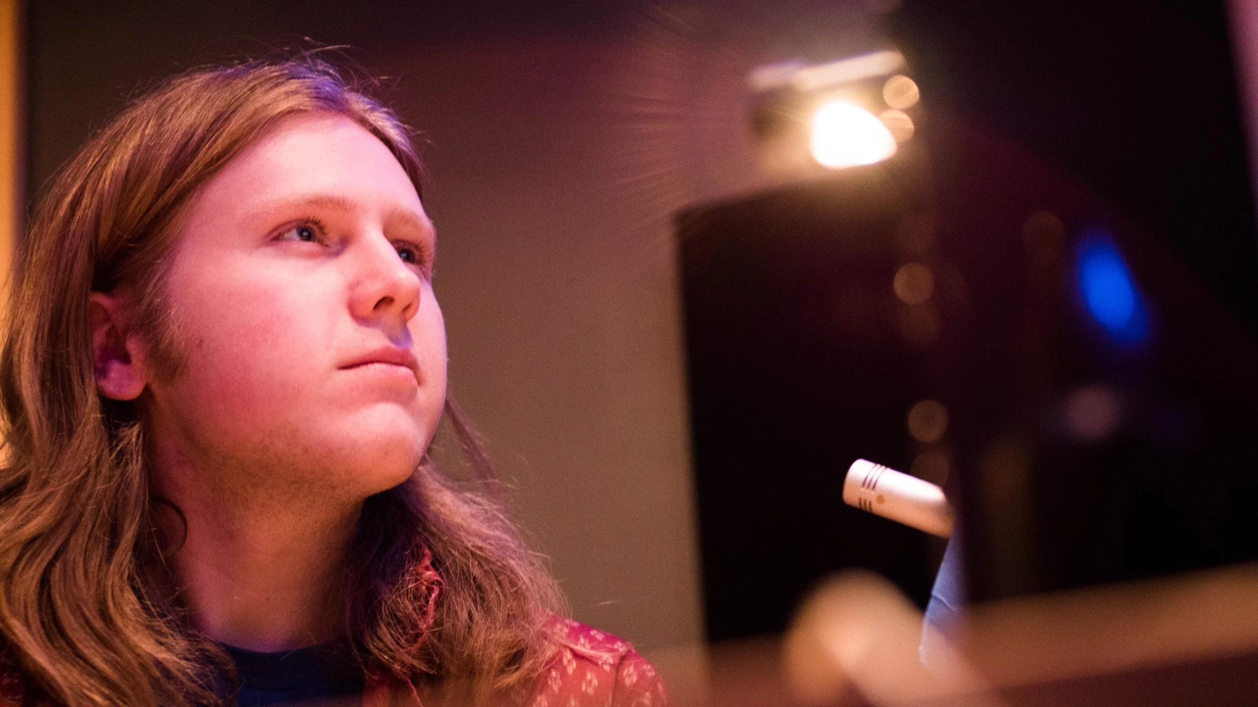 A young woman with long, wavy brown hair looking upward, possibly practicing singing or speaking, with a blurred microphone in front of her and a dark background with bright lo light.