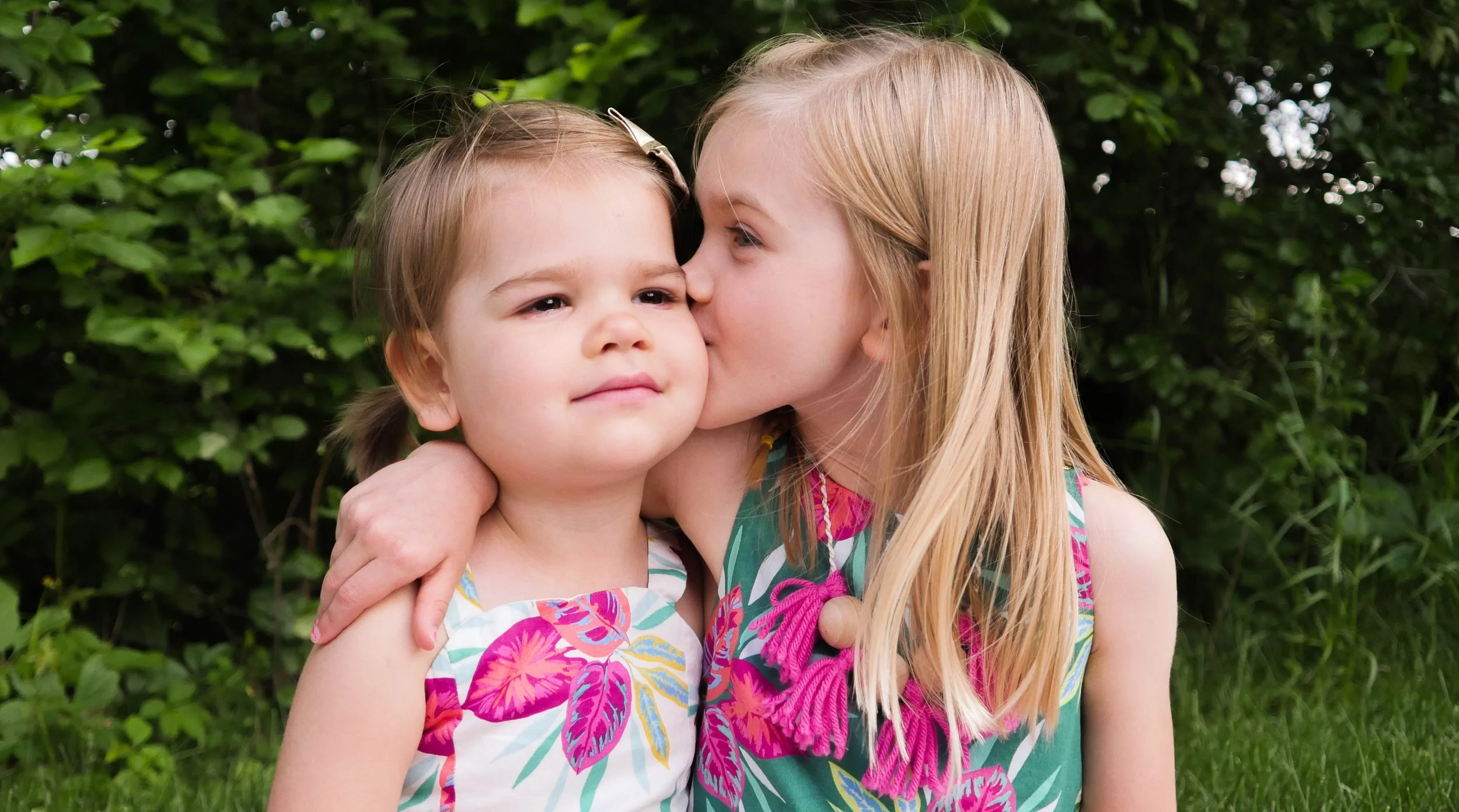 A young girl with long blonde hair and a girl with brown hair hugging outdoors in front of green foliage. The girl with blonde hair is kissing the brown-haired girl on the cheek.
