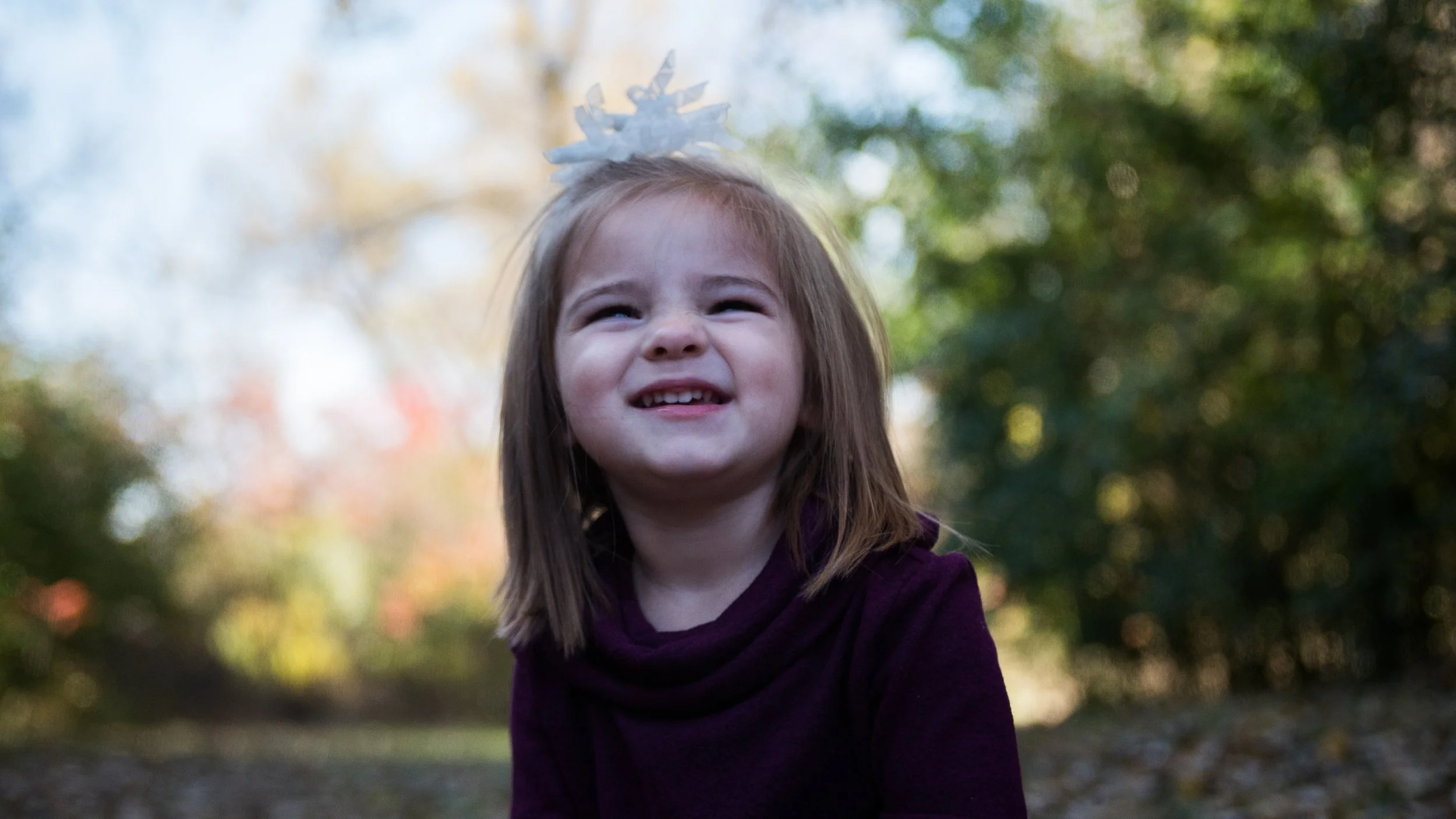 A young girl with brown hair smiling outdoors, wearing a dark purple shirt and a white hair accessory, with blurry trees and fall foliage in the background.