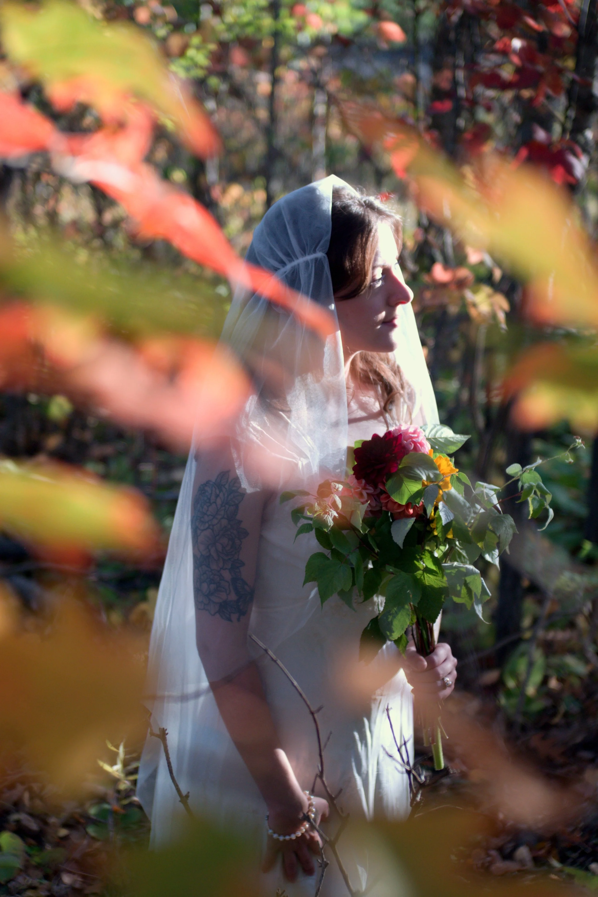 A woman in a white wedding dress and veil standing outdoors among autumn-colored leaves, holding a bouquet of flowers.