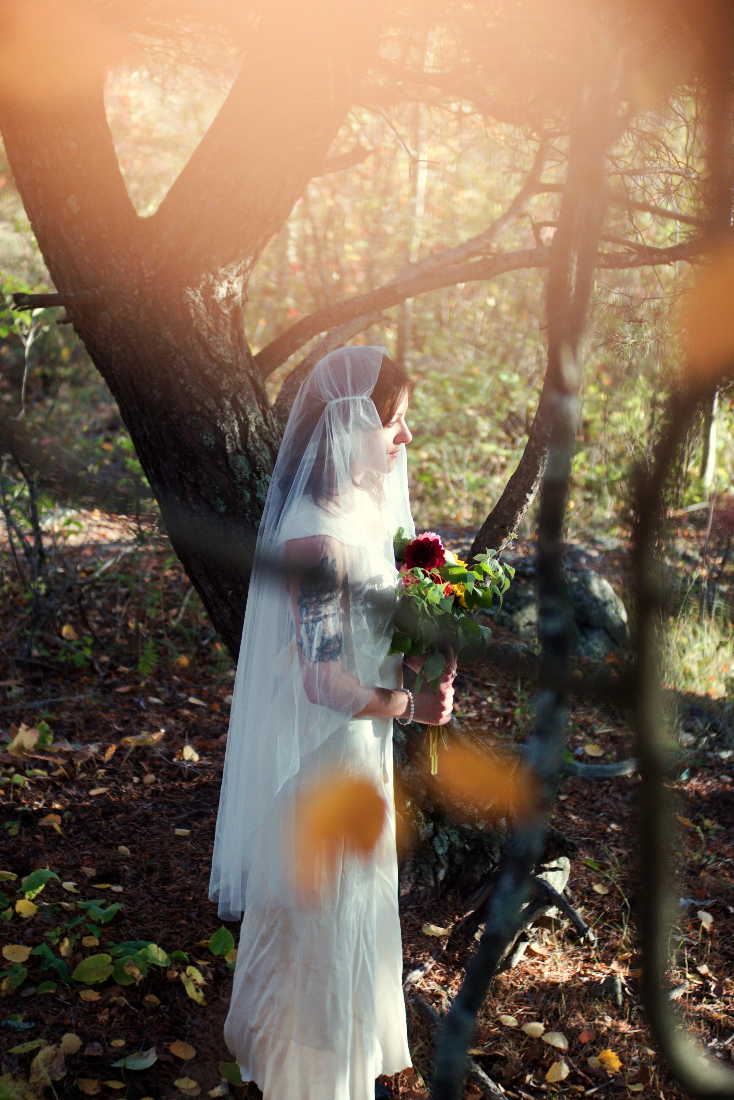 A woman wearing a white dress and a veil stands in a wooded area, holding a bouquet of flowers, with sunlight filtering through the trees.