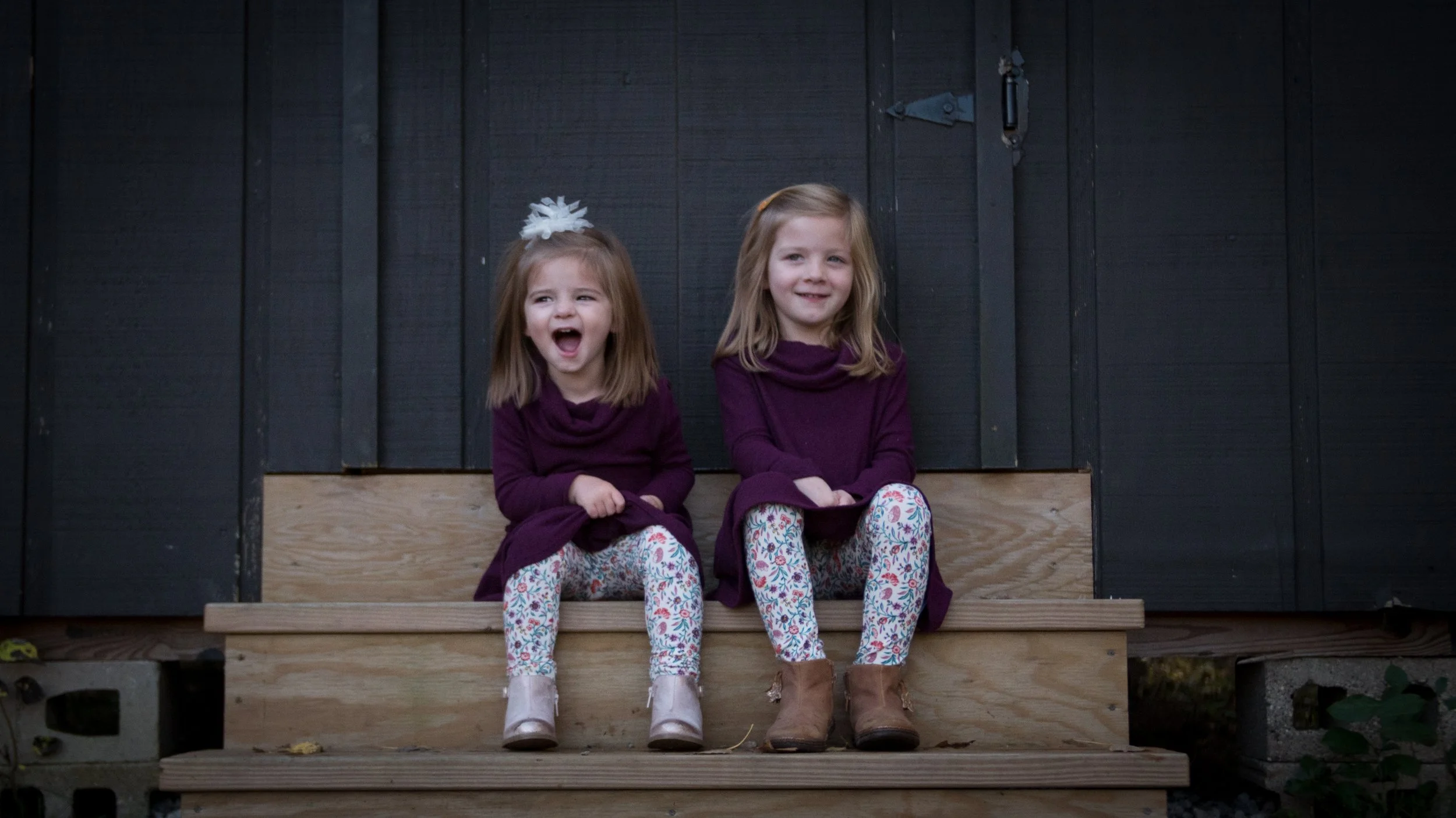 Two young girls sitting on wooden steps in front of a black wooden wall, smiling at the camera.