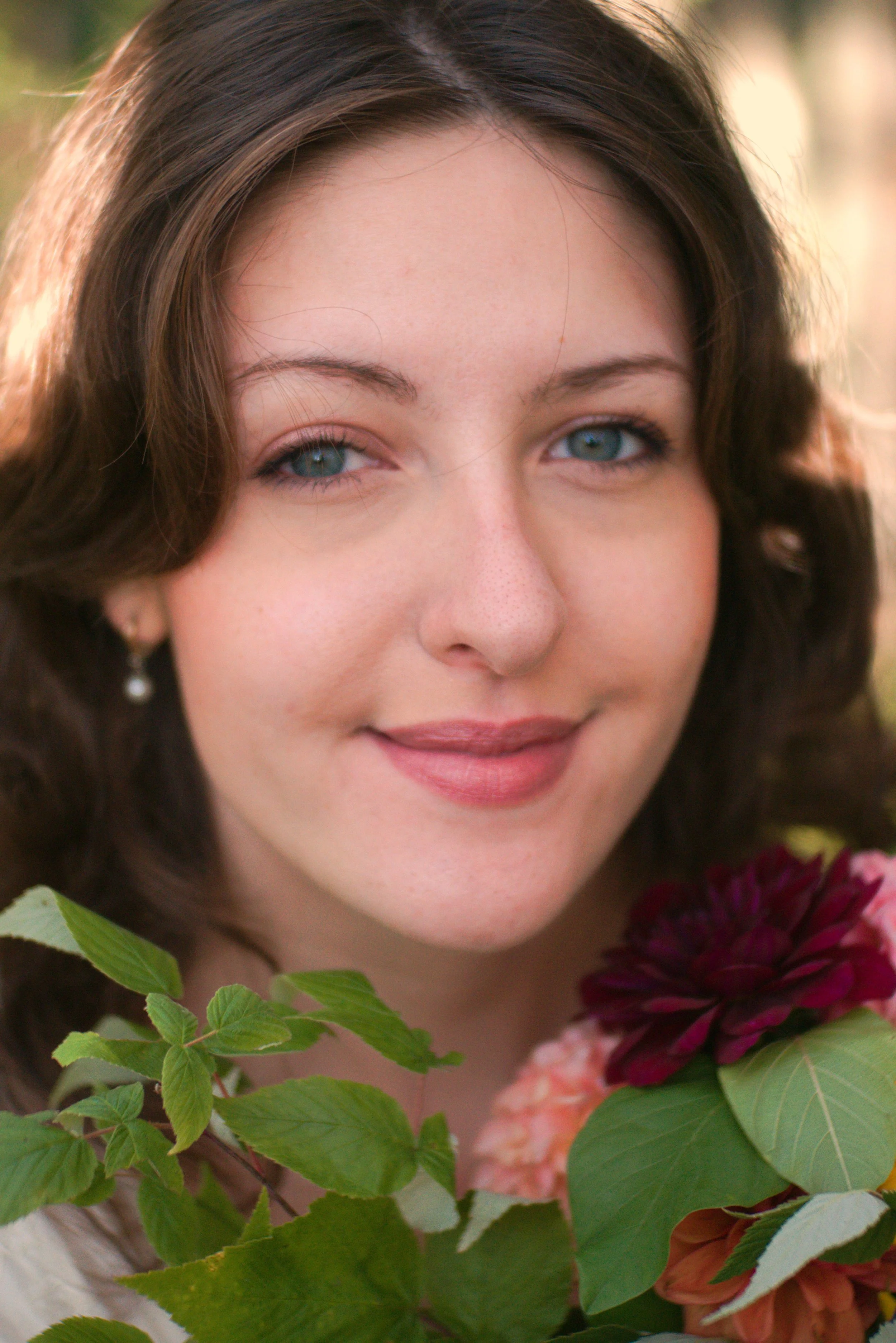 Close-up of a woman with brown hair and blue eyes, partially obscured by green leaves and burgundy flowers.