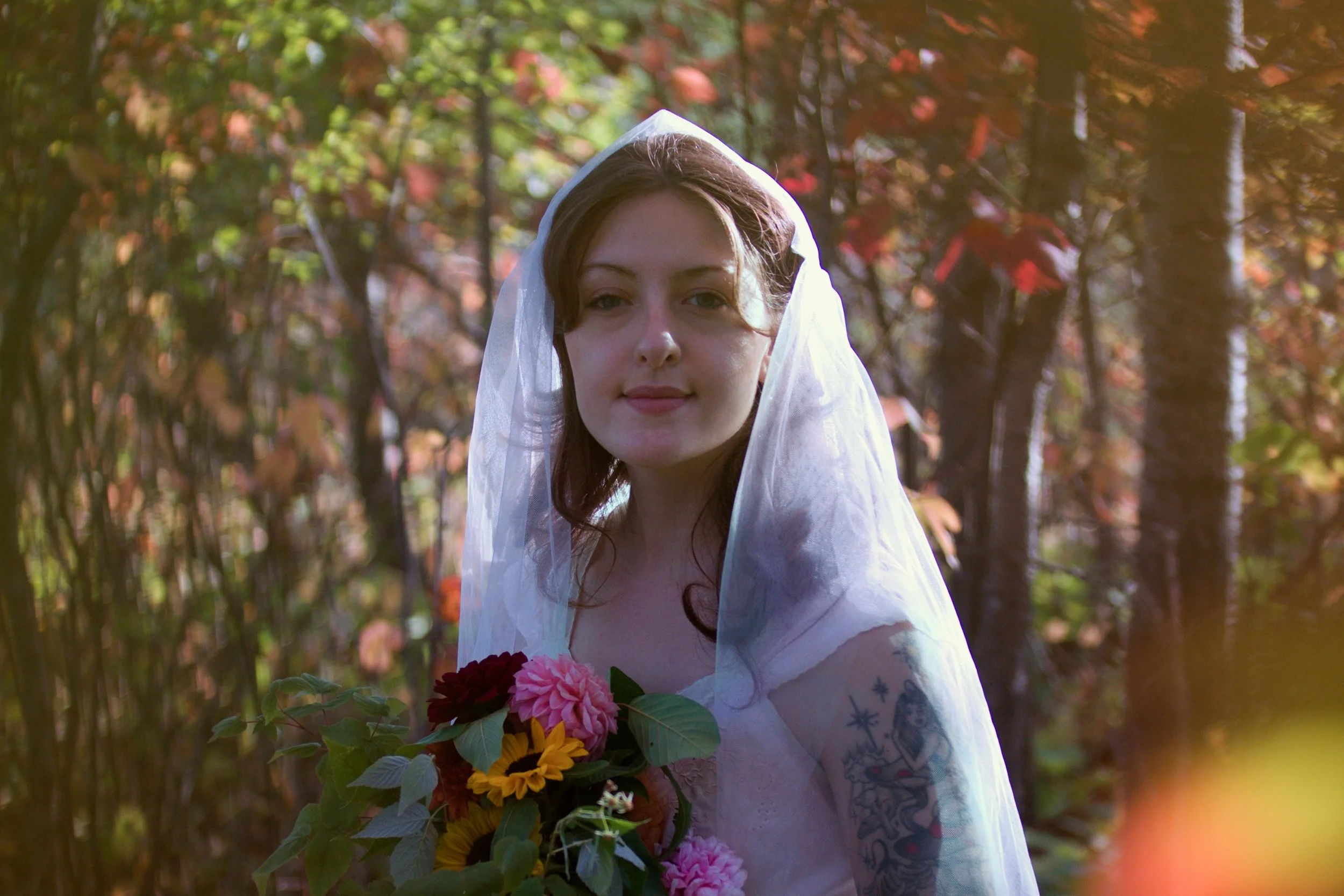 Young woman in a white veil holding a bouquet of colorful flowers in a wooded area with sunlight filtering through trees.