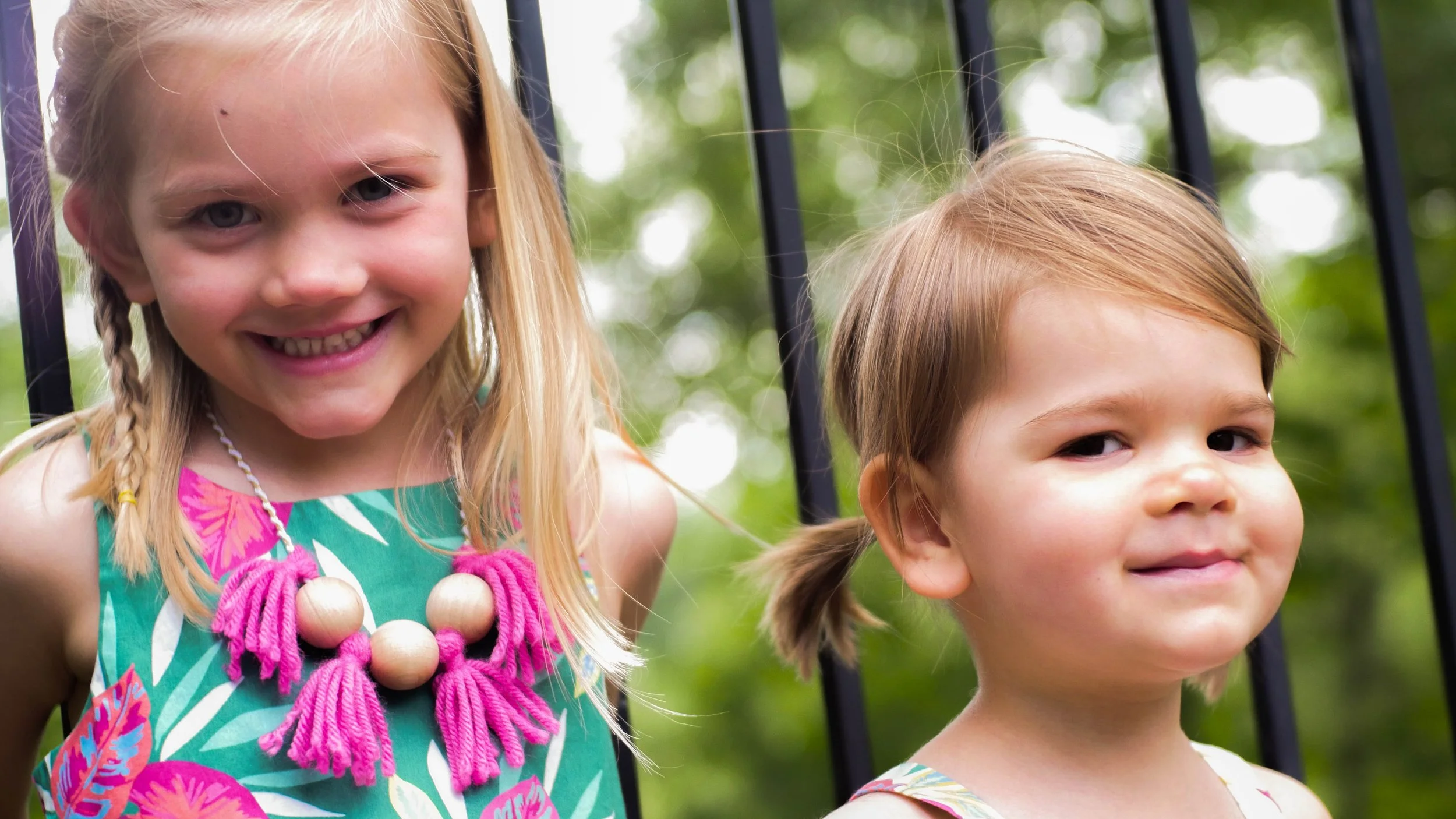Two young girls with blonde hair outdoors, one smiling and the other with a neutral expression, with green trees and a black metal fence in the background.