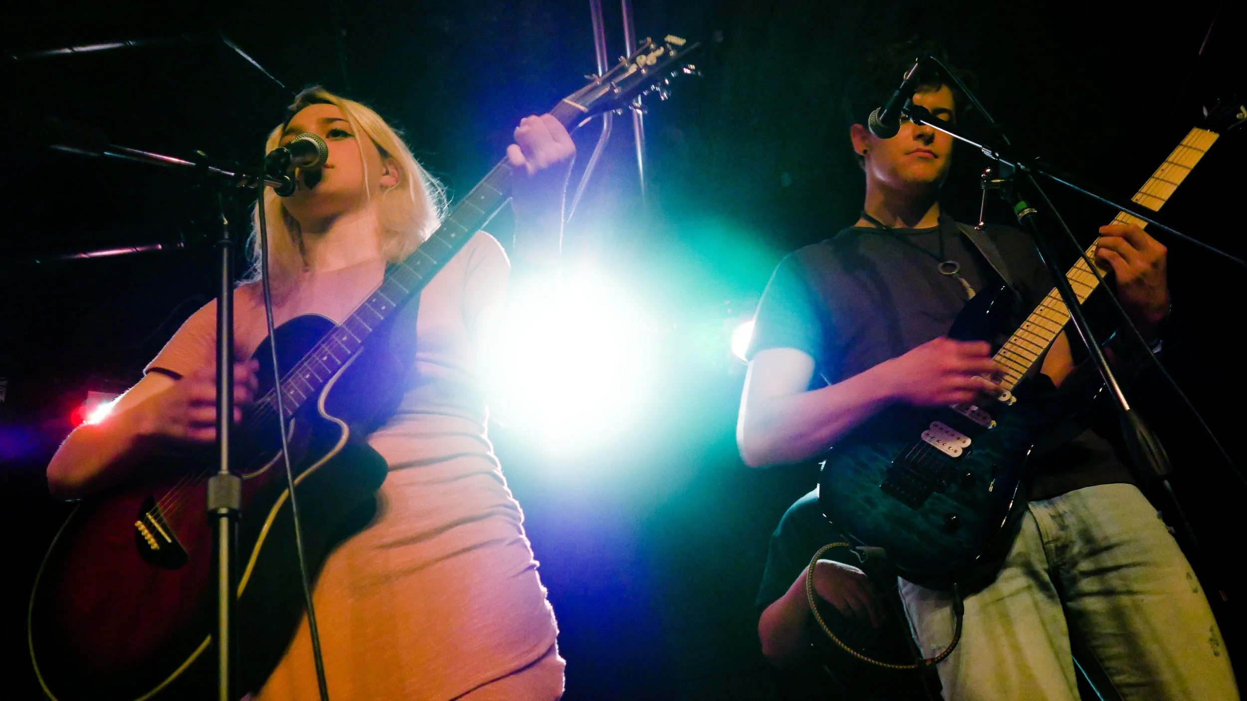 Two musicians, a woman and a man, performing on stage with guitars under stage lighting, microphones, and a dark background.