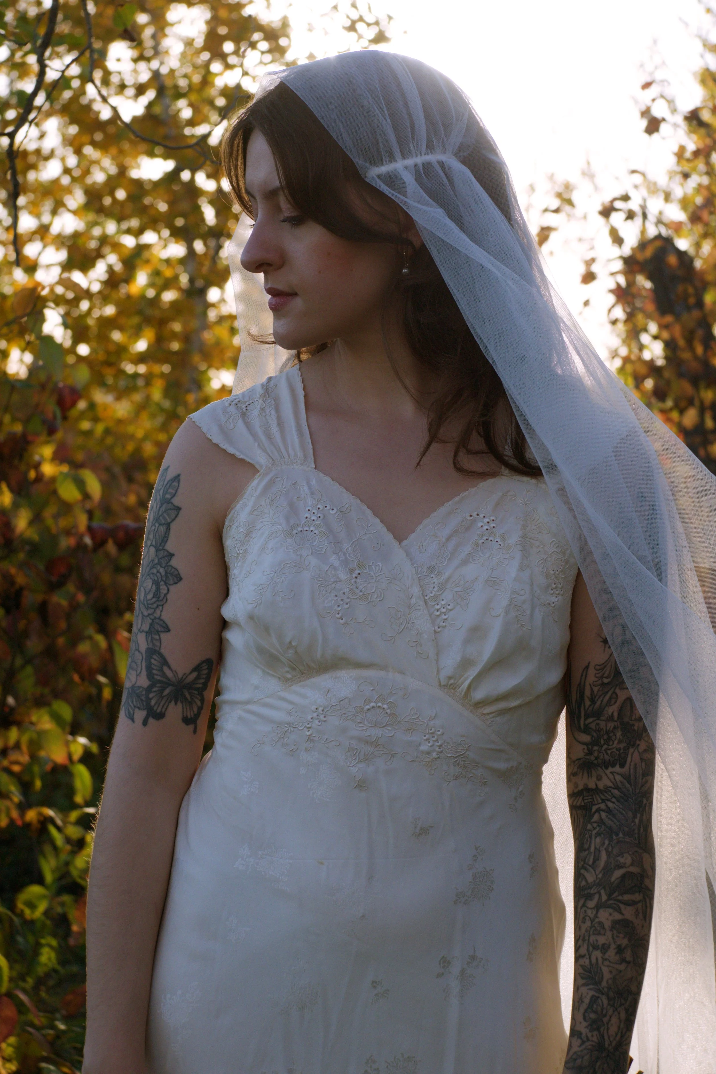 A woman wearing a wedding dress and veil standing outdoors with trees and sunlight in the background.