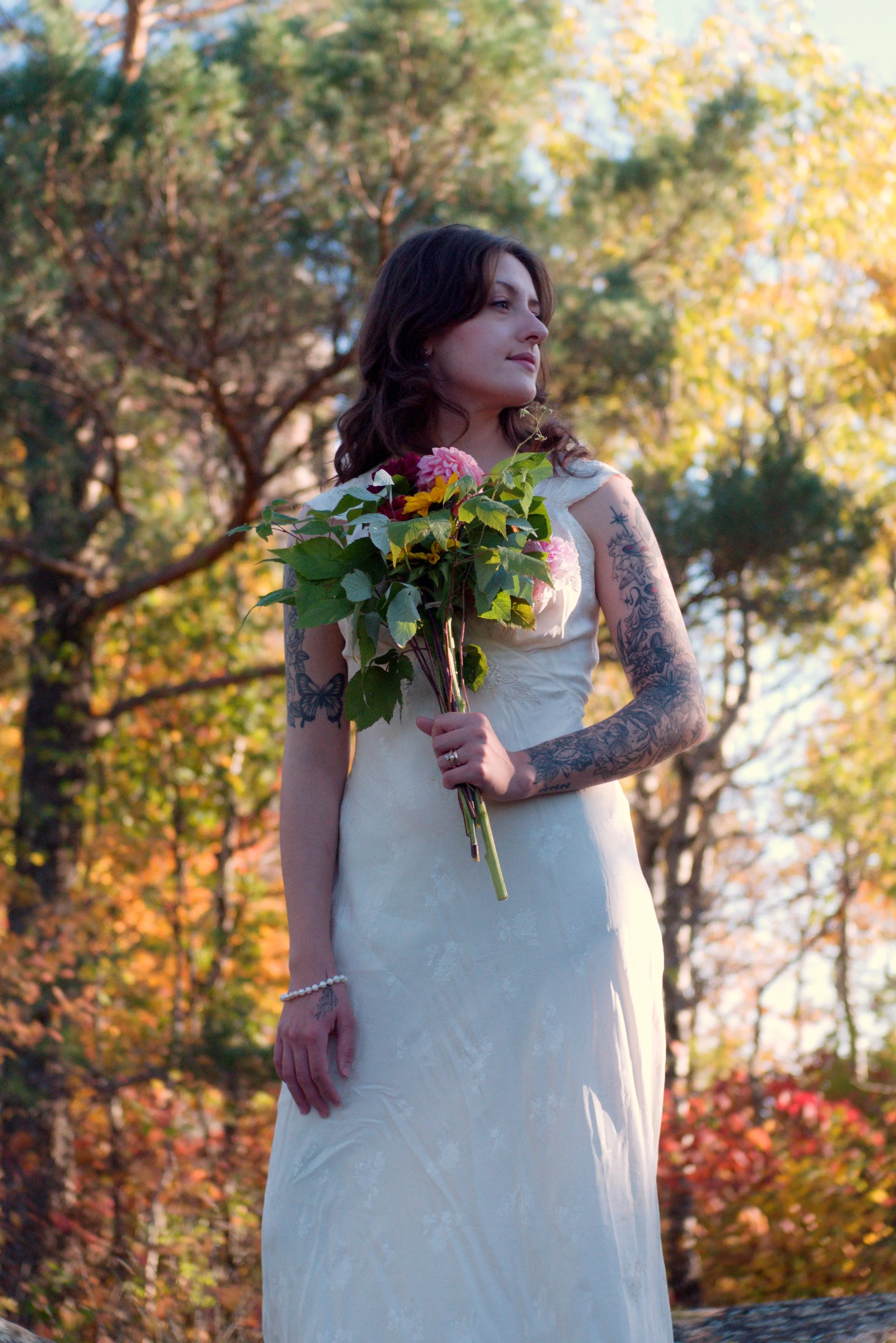 A woman with dark brown hair and tattoos on her arms, wearing a white dress, holding a bouquet of colorful flowers, standing outdoors in a park with trees and autumn leaves in the background.