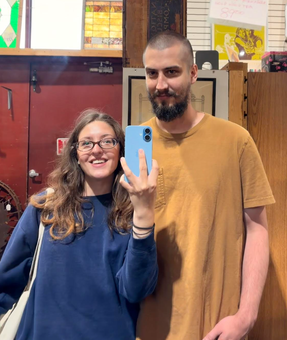 A young woman with glasses and brown hair taking a mirror selfie with a man who has a beard and is wearing a yellow t-shirt in a store or antique shop.