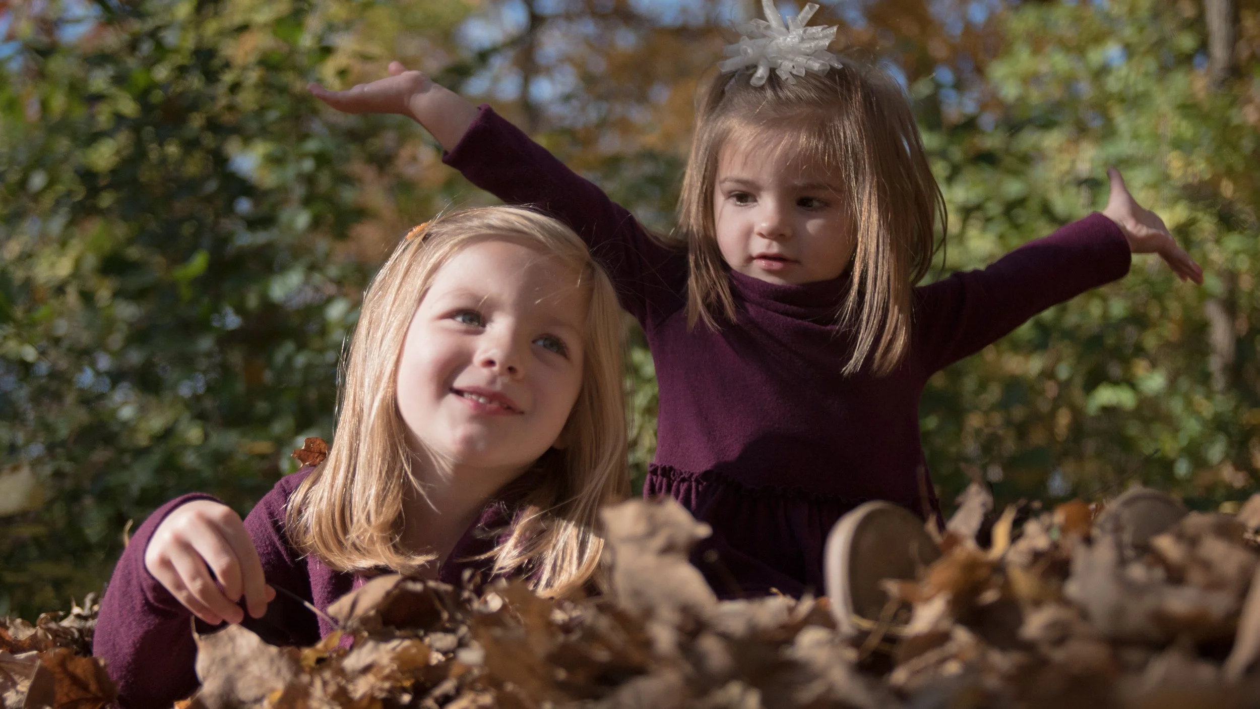 Two young girls playing in the autumn leaves outdoors. One girl is looking up with her arms outstretched, while the other girl is smiling and looking at the camera. Both are wearing maroon dresses and surrounded by trees with fall foliage.