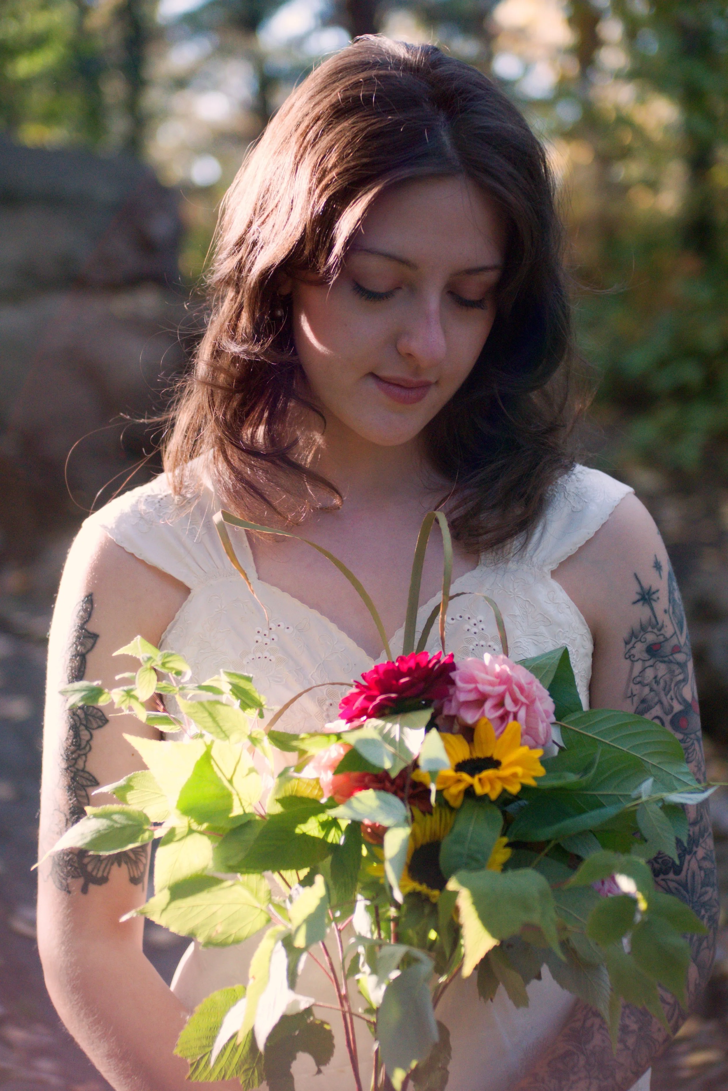 A young woman with dark brown hair and tattoos on her arm is holding a bouquet of colorful flowers, including pink, red, yellow, and orange blooms, outdoors with blurred trees in the background.