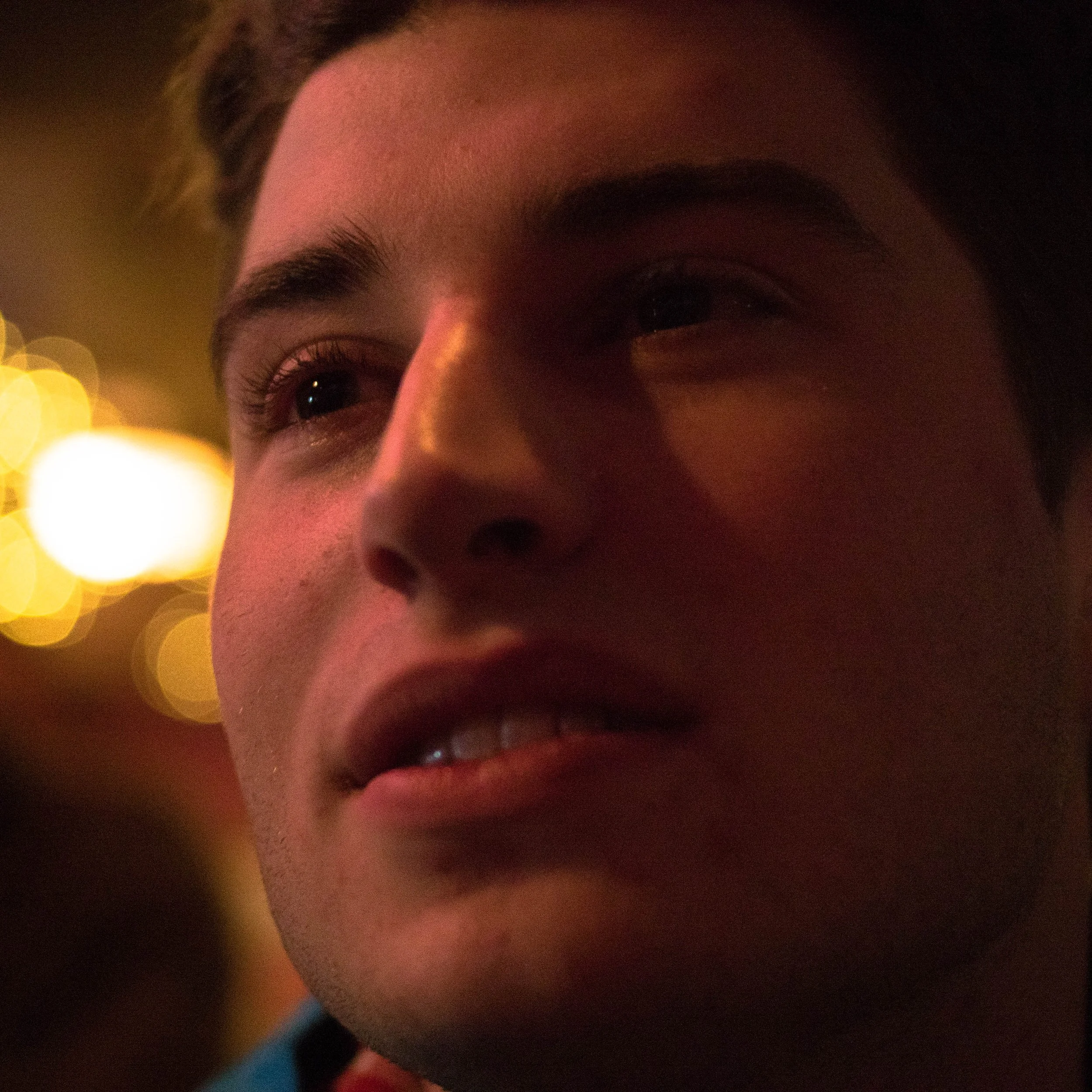 Close-up of a person's face showing partial view of eyes, eyebrows, and nose, with a warm, blurred background of lights.