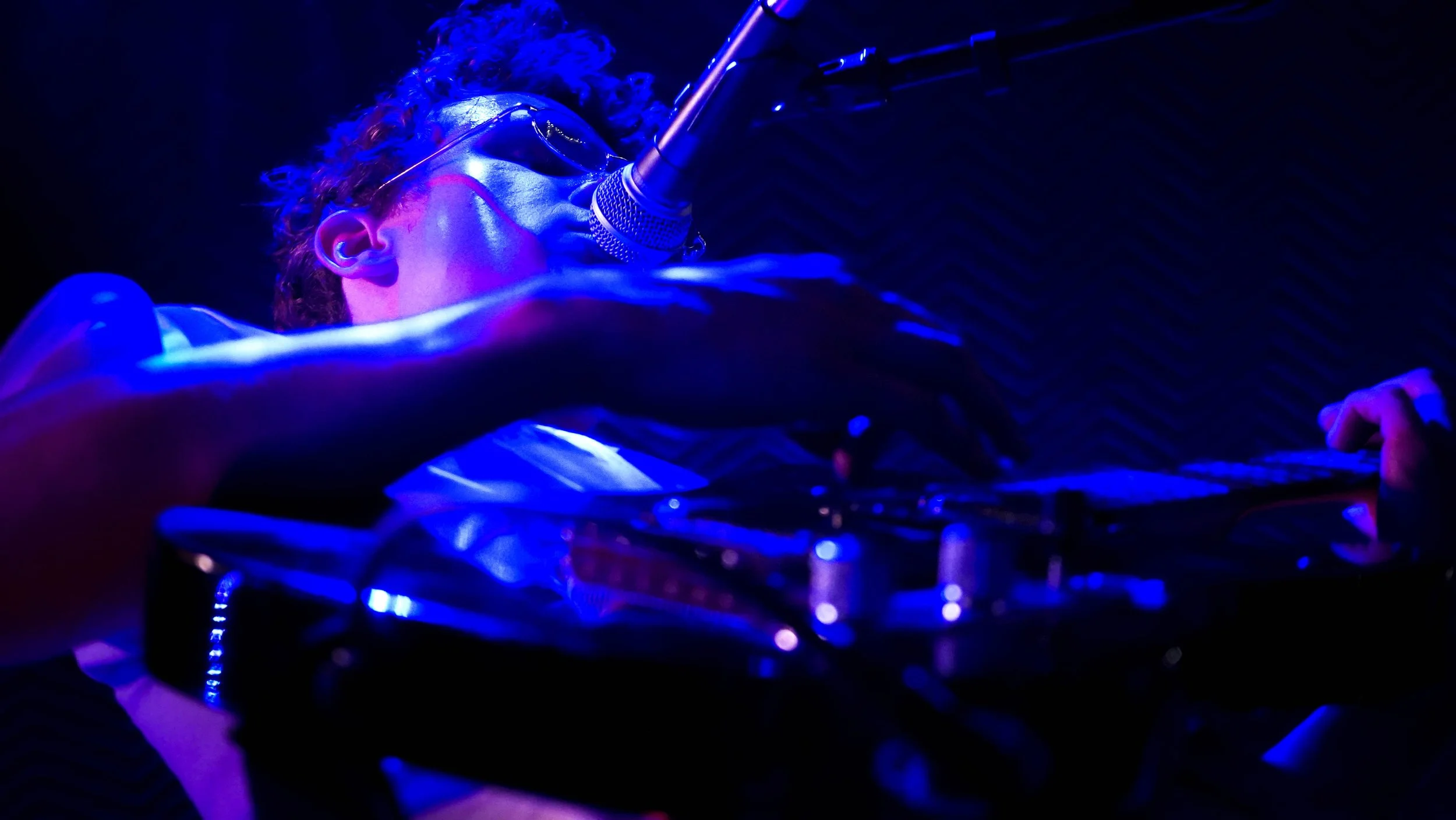 A person with curly hair and glasses singing into a microphone while playing a keyboard under blue stage lighting.