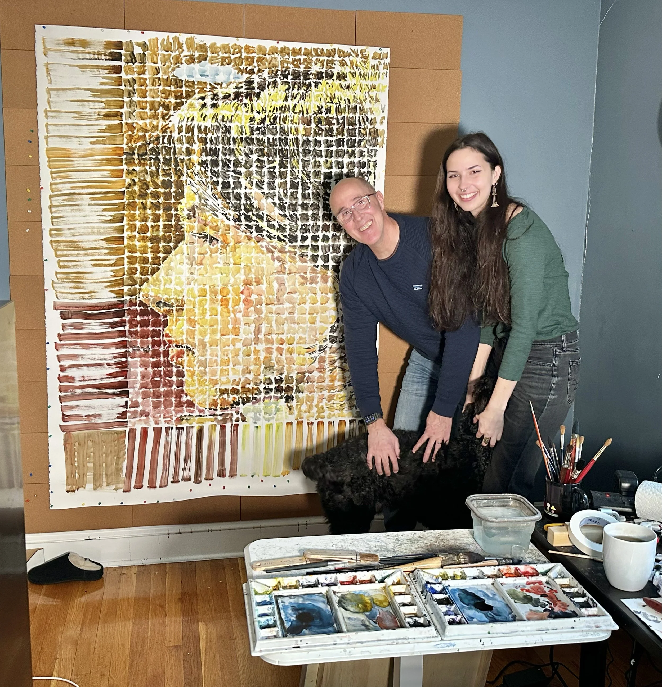 Two smiling people, a man and a woman, stand next to a large abstract painting while holding a black dog. The painting features colorful exaggerated portrait of a woman with glasses, created with broad brushstrokes. There are painting supplies and brushes on a nearby table.