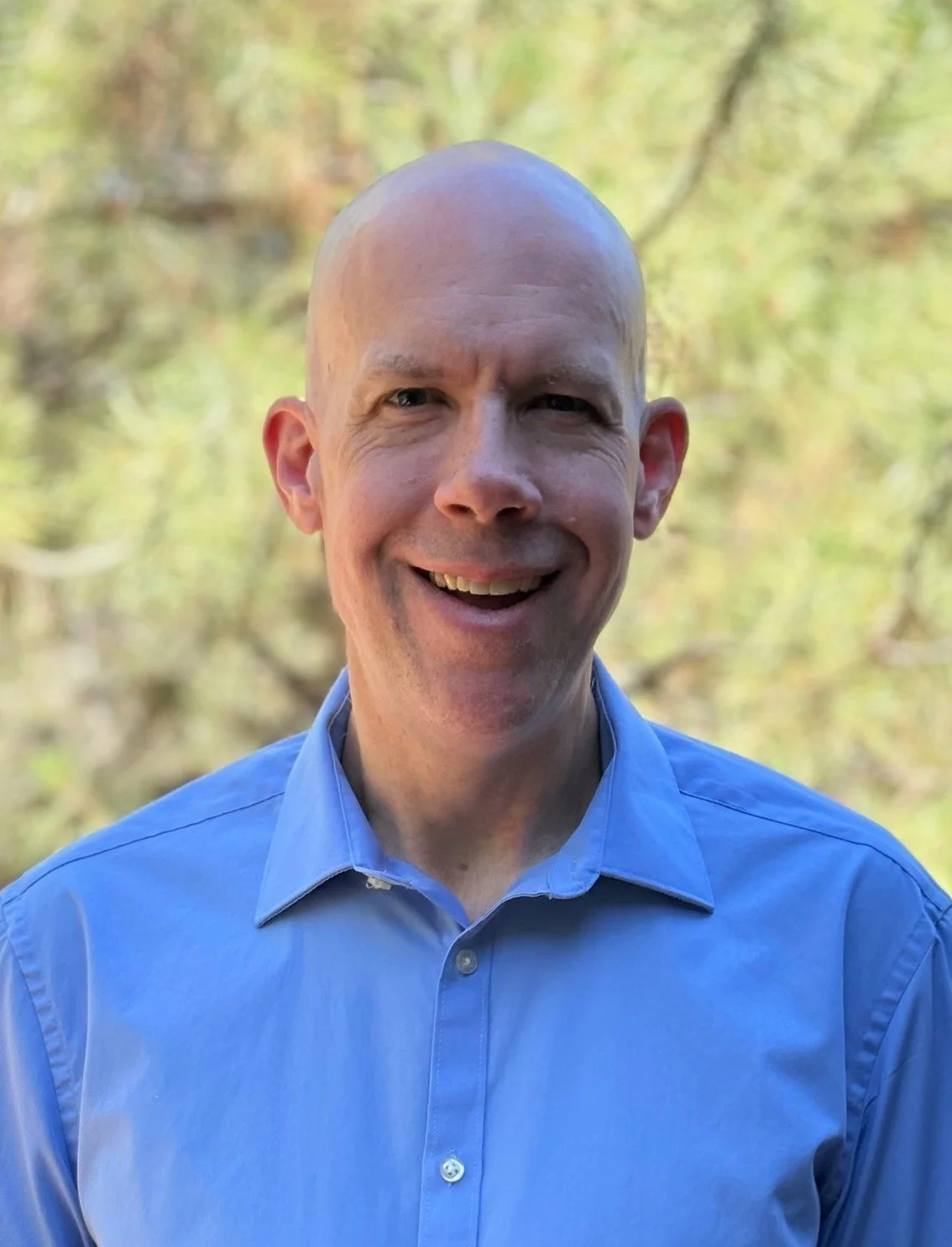 A smiling man wearing a blue button-up shirt outdoors with a blurred background of trees and greenery.