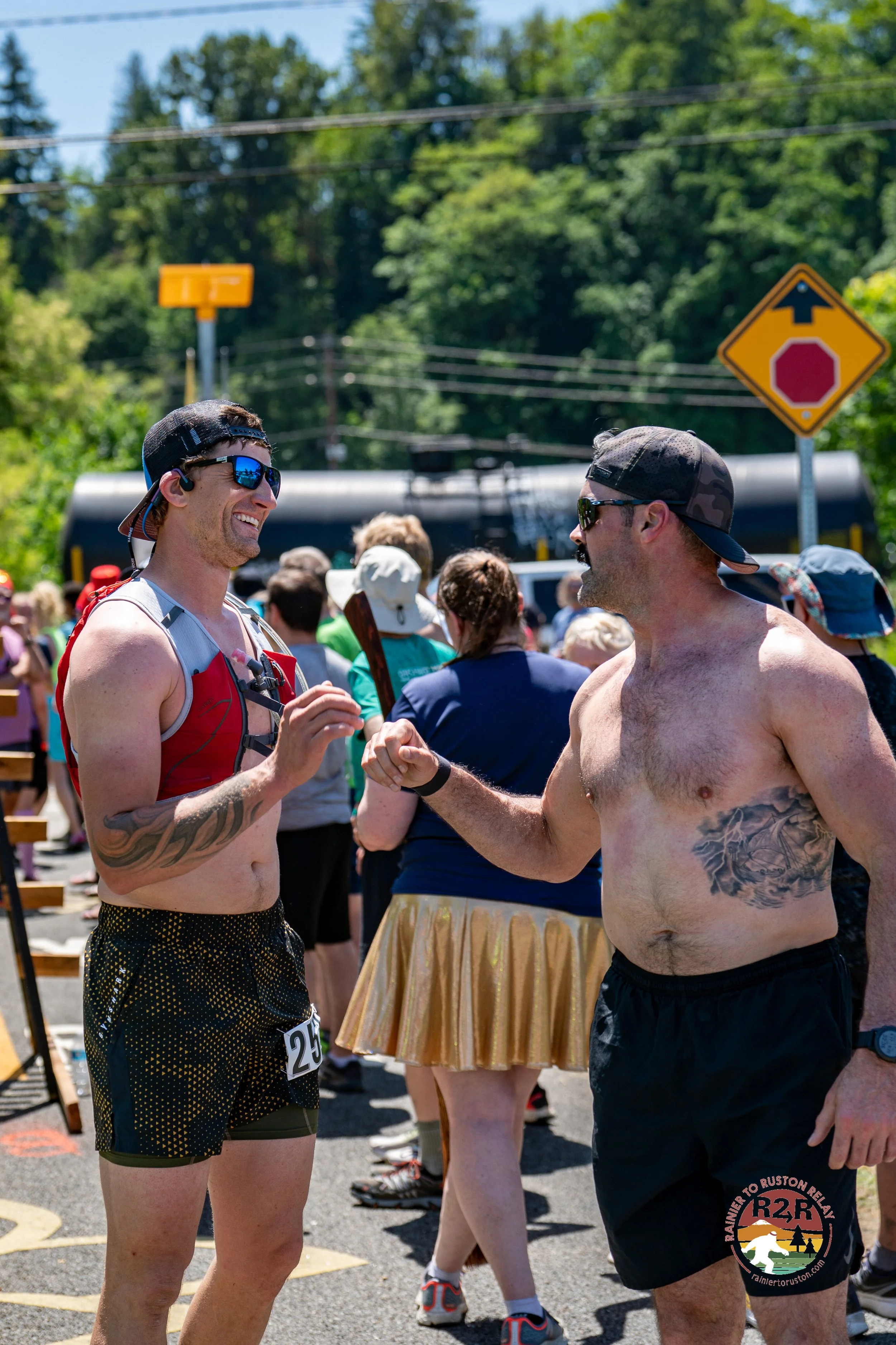 Two shirtless men wearing sunglasses shaking hands and smiling at an outdoor event, with a crowd of people and trees in the background.